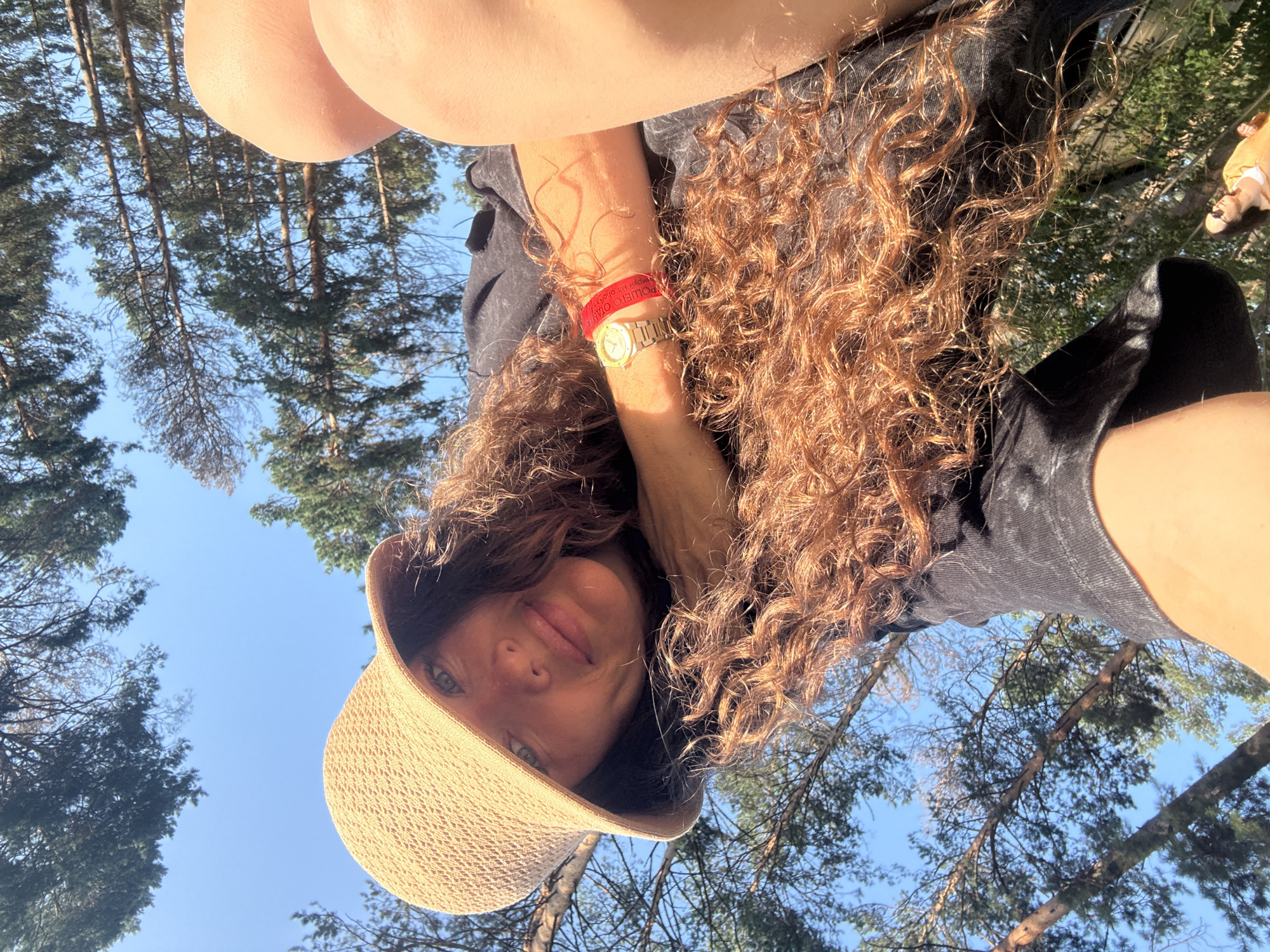 In a forest setting near the city of Barangol, Russia, a woman with long, curly brown hair and a wide-brimmed straw hat is captured in a close-up, low-angle selfie. She is wearing a black t-shirt and a red bracelet on her left wrist, which features some text. A watch with a gold band is also visible on the same wrist. The background consists of tall pine trees against a clear blue sky. In the lower-left corner of the frame, a second individual wearing a yellow jacket and sunglasses can be seen walking away from the camera. The scene appears to be an outdoor recreational or travel setting.