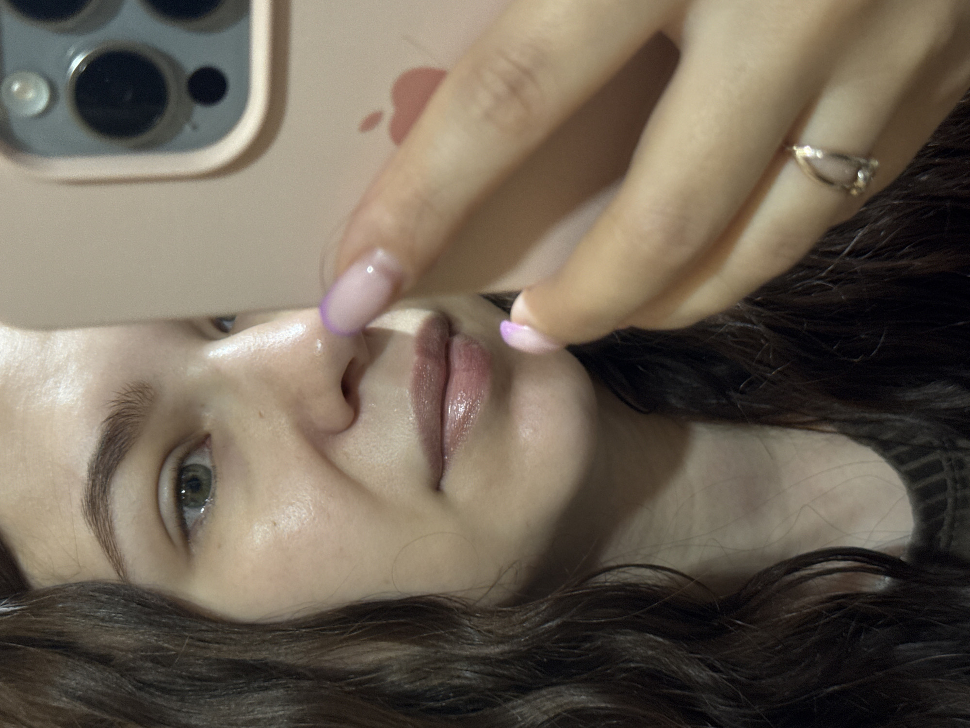 A close-up, slightly rotated photograph shows a woman's face as she takes a selfie. The left side of her face is visible, including her eye, nose, and lips. She has dark eyebrows and light-colored eyes. She is holding a pink smartphone, likely an iPhone, with its camera lenses positioned above her eye. Her hand is visible, wearing a ring on one of her fingers. Her nails are painted with a light pink color and a purple tip. Her dark curly hair frames the lower part of her face. The environment is not visible; therefore, determining if the location is in Nizhnyaya Gar', Russia, is impossible.