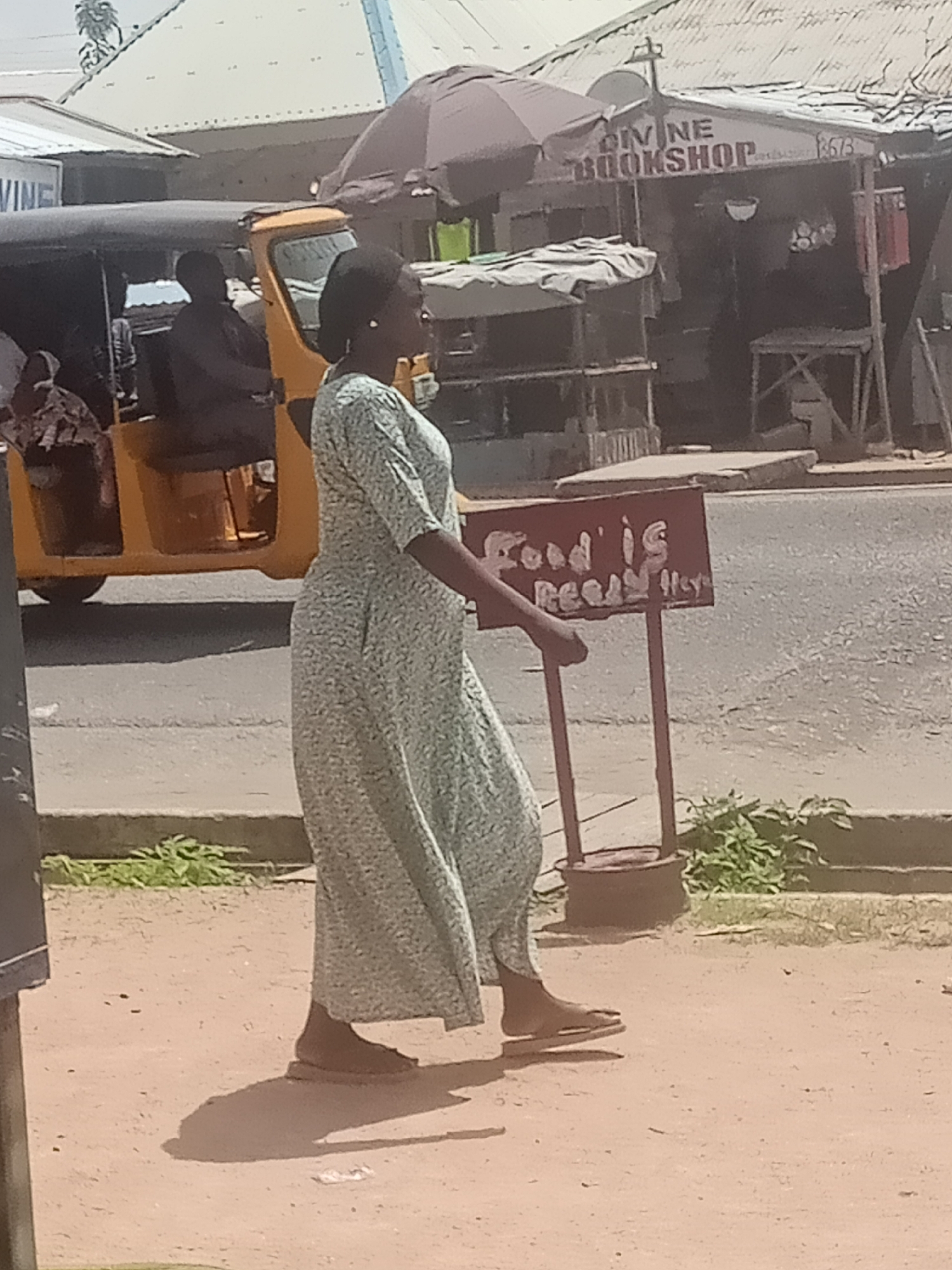 A woman in a light-colored, floral-patterned maxi dress walks on a dusty, unpaved sidewalk next to a paved road in Jalingo, Nigeria. The woman is facing to the right, with her left arm slightly extended, holding a red sign with white lettering that reads "Food is Ready". She is wearing flat sandals. Behind her, a yellow motorized tricycle, commonly known as a "Keke Napep", is parked. In the background, a shop with a corrugated metal roof displays a sign that reads "DIVINE BOOKSHOP". Various goods are visible on shelves and tables outside the shop, and there are awnings over some of the structures. The environment appears to be an urban street scene with a mix of commercial activity and unpaved areas.