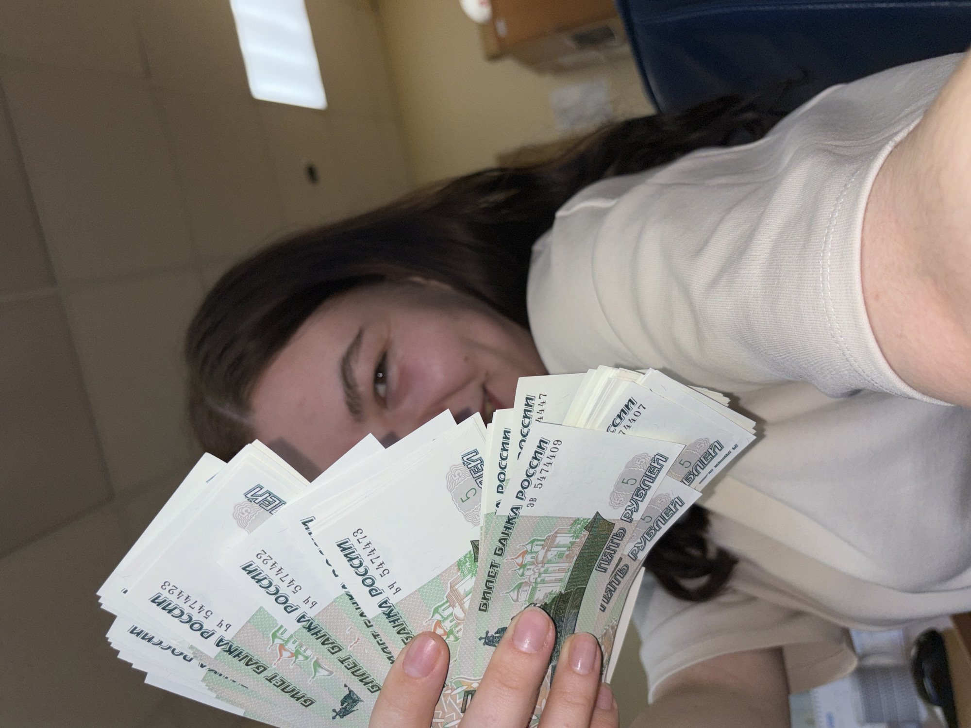 A woman is holding a stack of Russian 5-ruble bills. The bills are green and white with Cyrillic text and images of buildings. She is smiling slightly and wearing a white t-shirt. The setting is indoors with neutral wall color and possible desk visible. There is no information available about her location in Perm, Russia.