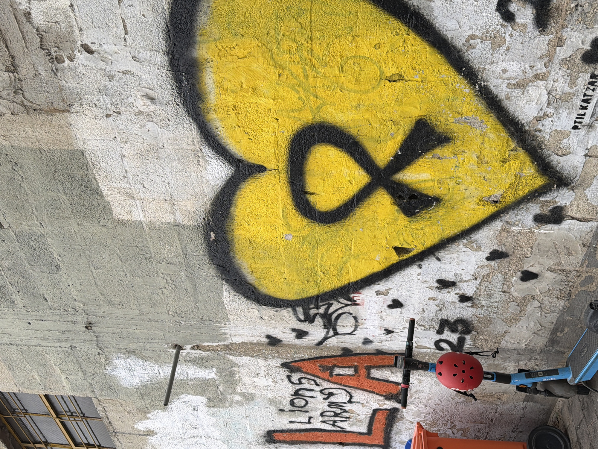 The image depicts a segment of a wall in an urban environment, possibly located in Tel-Aviv, Israel. The wall is covered with graffiti, including a large yellow heart with a black outline and an infinity symbol inside. Other graffiti includes the words "LIONS ARMY" in orange and black, along with other indistinct markings. A blue scooter is leaning against the wall, with a red helmet placed on the handlebars. The wall appears to be made of concrete and shows signs of weathering and discoloration. Part of a window with grilles is visible at the bottom-left.
