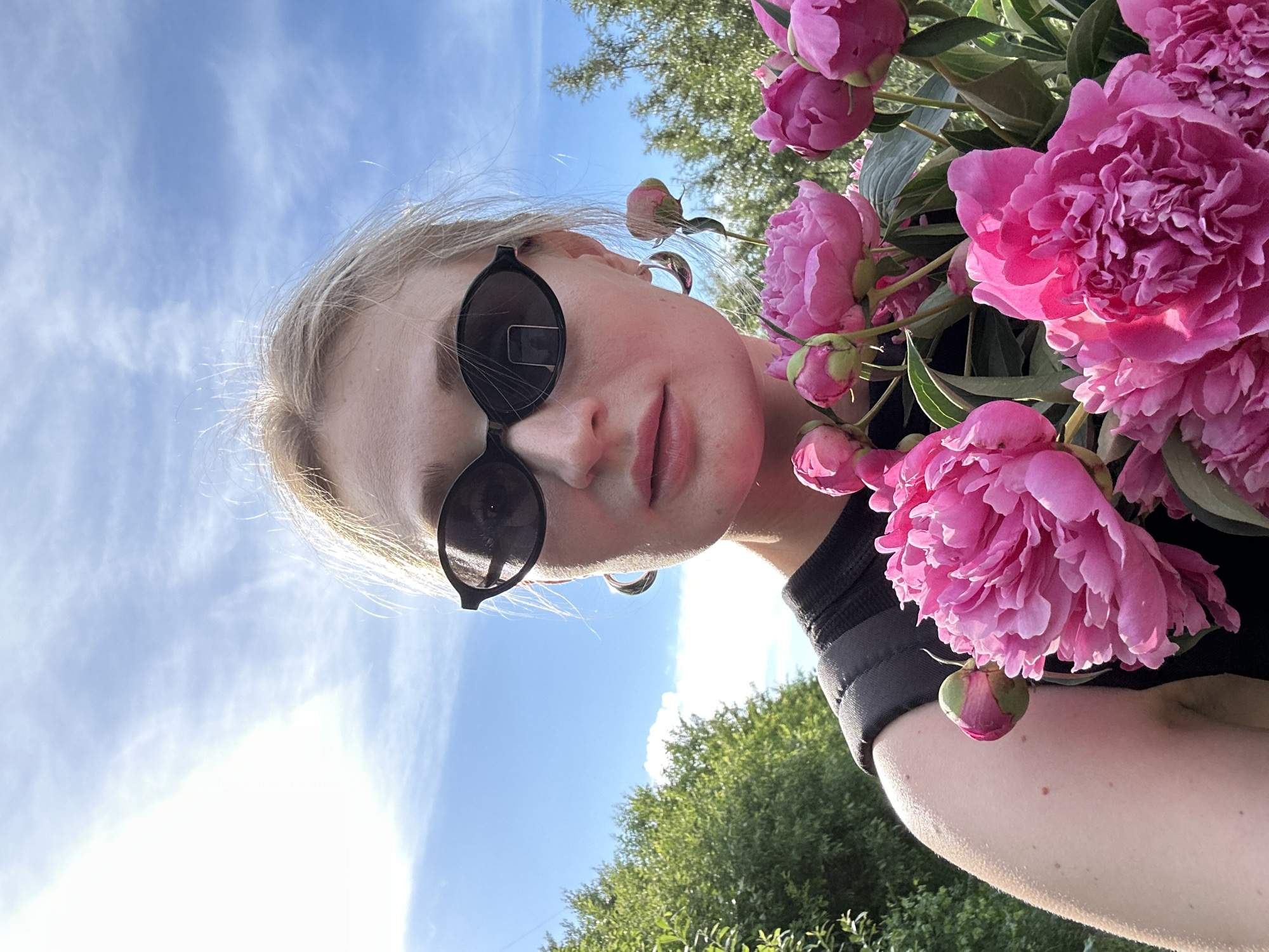 This is a selfie of a young Caucasian woman with blonde hair and sunglasses, holding a bouquet of pink peonies. She is outdoors, with a blue sky and green trees in the background. The woman is wearing a black top and large hoop earrings. The overall lighting suggests it is daytime, likely sunny. The scene is set in Chaikovsky, Russia, as indicated by the provided context.