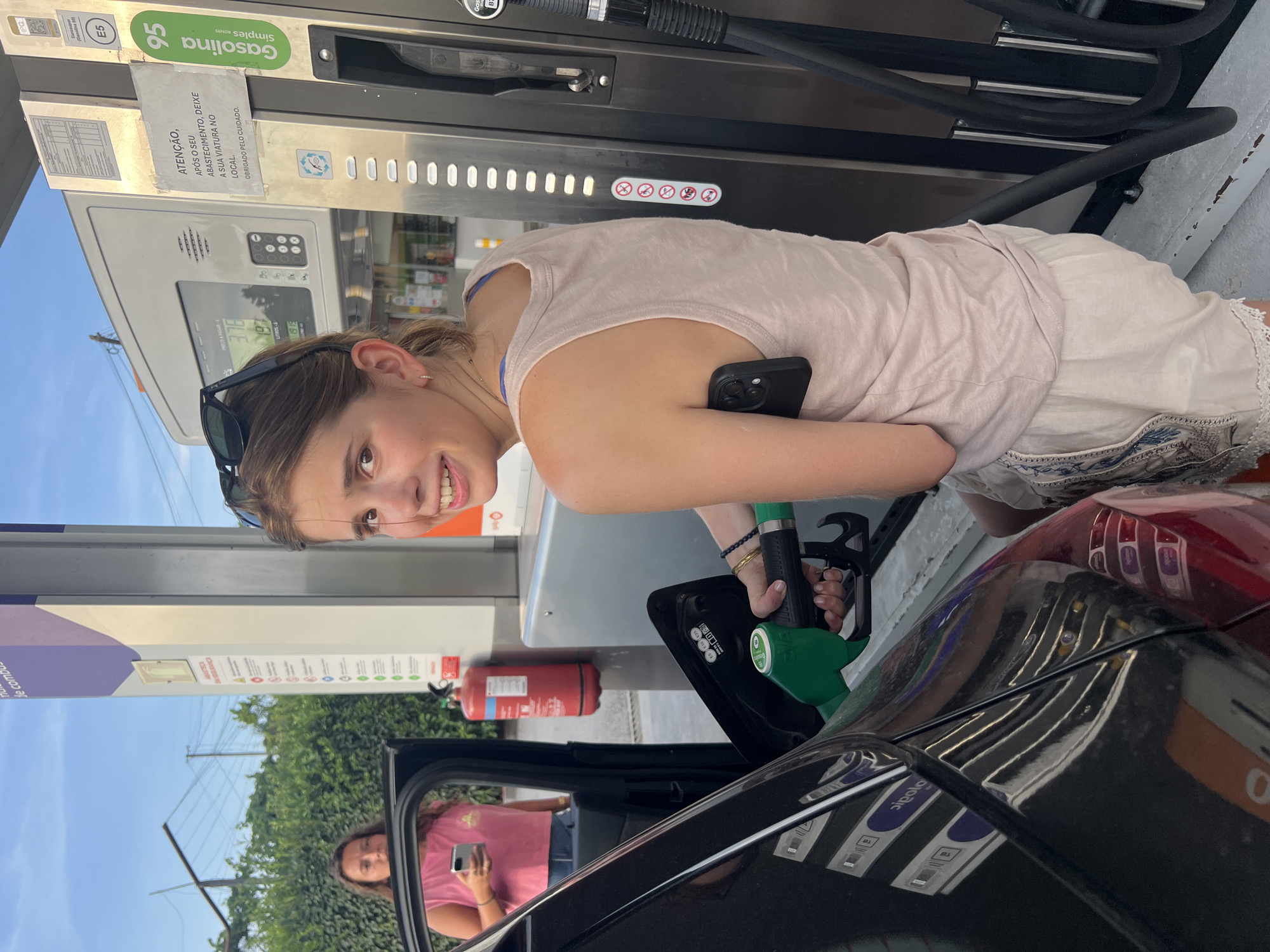 An outdoor medium shot shows a young woman with light brown hair and a surprised expression, looking over her left shoulder at the camera. She is standing at a gas station in Felgueiras, Portugal, with the nozzle of a green gas pump in her right hand, connected to a black car. Her left arm is bent, and a black smartphone is tucked under her left armpit, protruding from her light pink tank top. The gas pump is tall and silver with a digital display showing numbers. Behind her, another young woman in a bright pink tank top is visible, standing next to the open car door and holding a smartphone. The background includes a gas pump sign with the number 95, a fire extinguisher, and trees. The lighting suggests it is late afternoon or early evening.