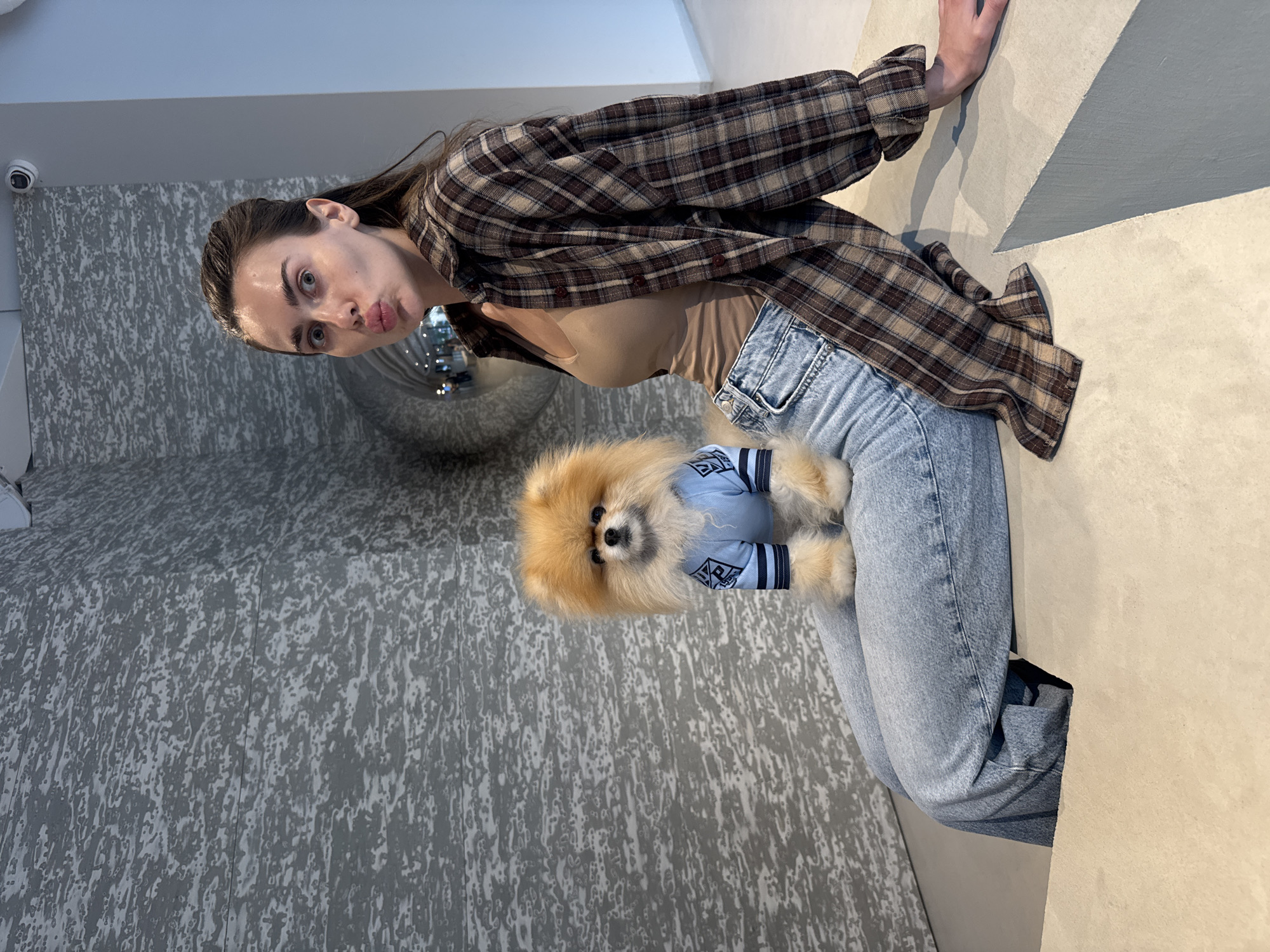A young woman is depicted sitting indoors, holding a small Pomeranian dog on her lap. She has long dark hair and is wearing a brown and beige plaid button-up shirt over a light beige top, paired with light blue jeans. Her lips are puckered in a playful expression as she looks directly at the camera.
The fluffy, light brown Pomeranian, positioned on her lap, is wearing a light blue sweater adorned with dark blue stripes and a white stylized pattern or text, which is not clearly legible. The dog is also looking towards the camera.
The setting appears to be a modern interior space. The floor is a light, neutral beige color, transitioning to a grey, textured wall with a mottled or streaky pattern. Another wall is a plain light beige. In the background, a reflective surface, possibly a mirror or polished metal, reflects some ambient lights. A small white security camera is mounted high on the textured grey wall. The overall mood of the image is affectionate and playful, capturing a moment between a pet owner and her dog.