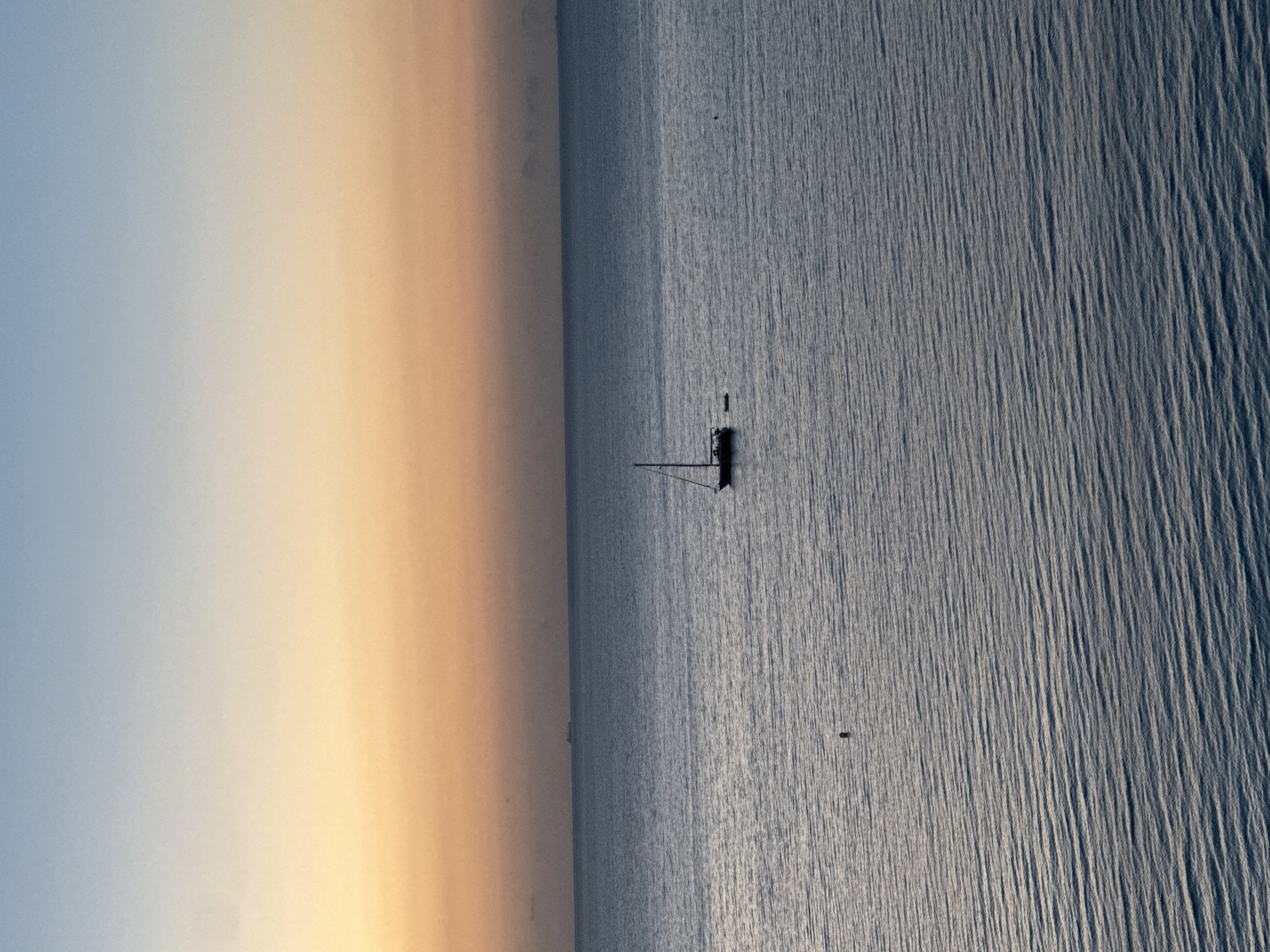 The image depicts a tranquil seascape at dawn or dusk, with a gradient of colors in the sky ranging from soft blue at the top to a warm orange and yellow near the horizon. The sea is calm, with gentle ripples reflecting the sky's colors. In the middle of the frame, a lone sailboat with its mast visible is anchored on the water. Further in the distance, a large cargo ship is barely visible on the horizon. A small dark buoy or marker floats in the water to the left of the sailboat. The scene suggests a serene coastal environment. While the image is evocative of coastal scenes, there is no specific indication of the location being Calp, Spain.