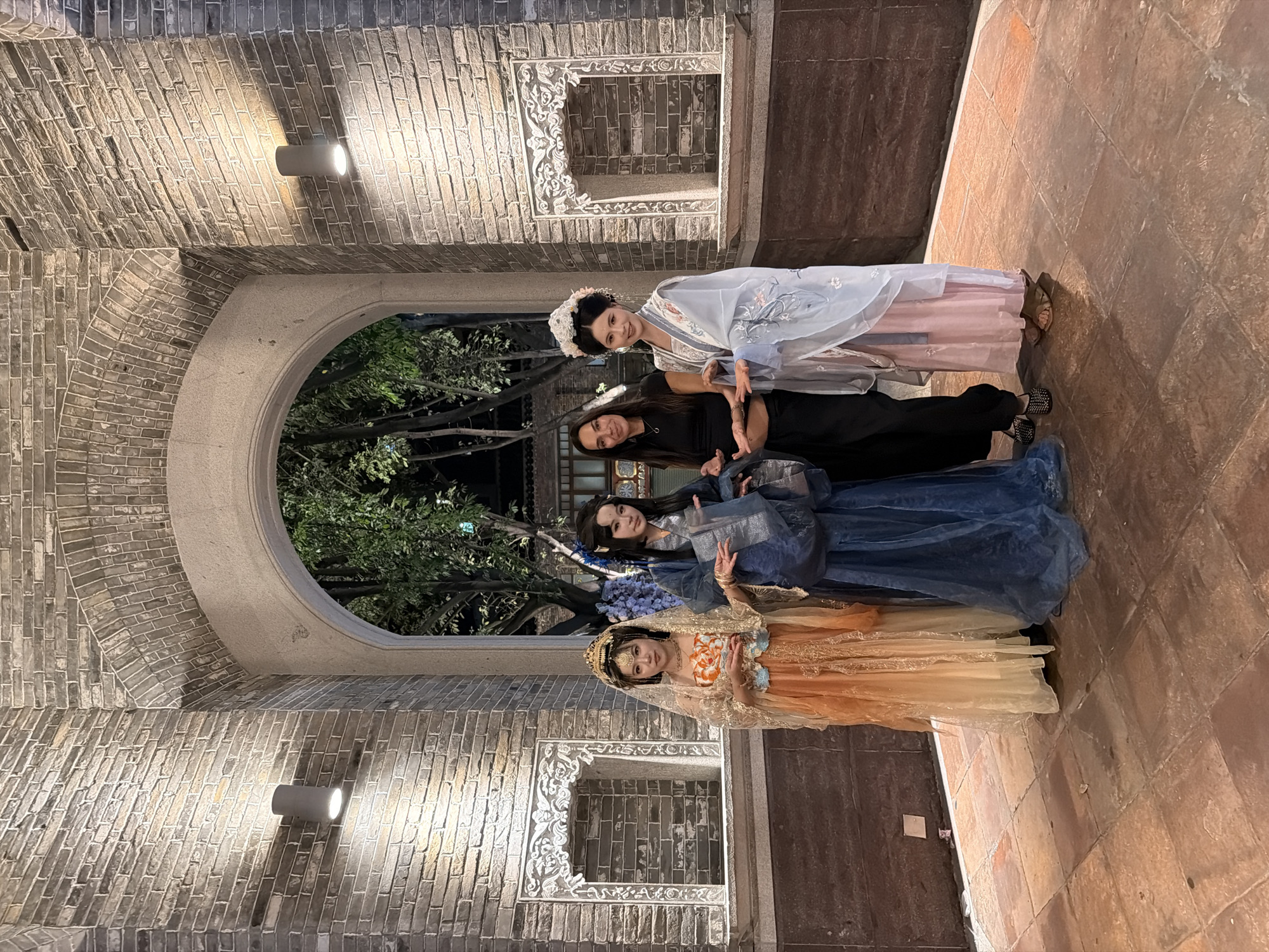 In the city of Chancheng District, China, four individuals stand within an archway constructed of gray brick. Two women on the left are dressed in traditional Chinese attire. The first woman, on the far left, wears an orange and gold layered gown with a sheer veil over her head, adorned with gold jewelry and a golden headpiece. The second woman, next to her, is in a flowing blue robe, her hair styled with blue accessories. The third individual, a woman in the center, wears a black short-sleeved top and black pants. The fourth individual, on the far right, is dressed in a light blue and pink traditional Chinese outfit, complete with a floral headpiece. The archway opens to a dimly lit area with trees visible in the background. The walls surrounding the archway are also made of gray brick, with decorative carved alcoves on either side. Two cylindrical wall lights are mounted on the left and right walls, casting a warm glow. The ground is paved with square, reddish-brown tiles.