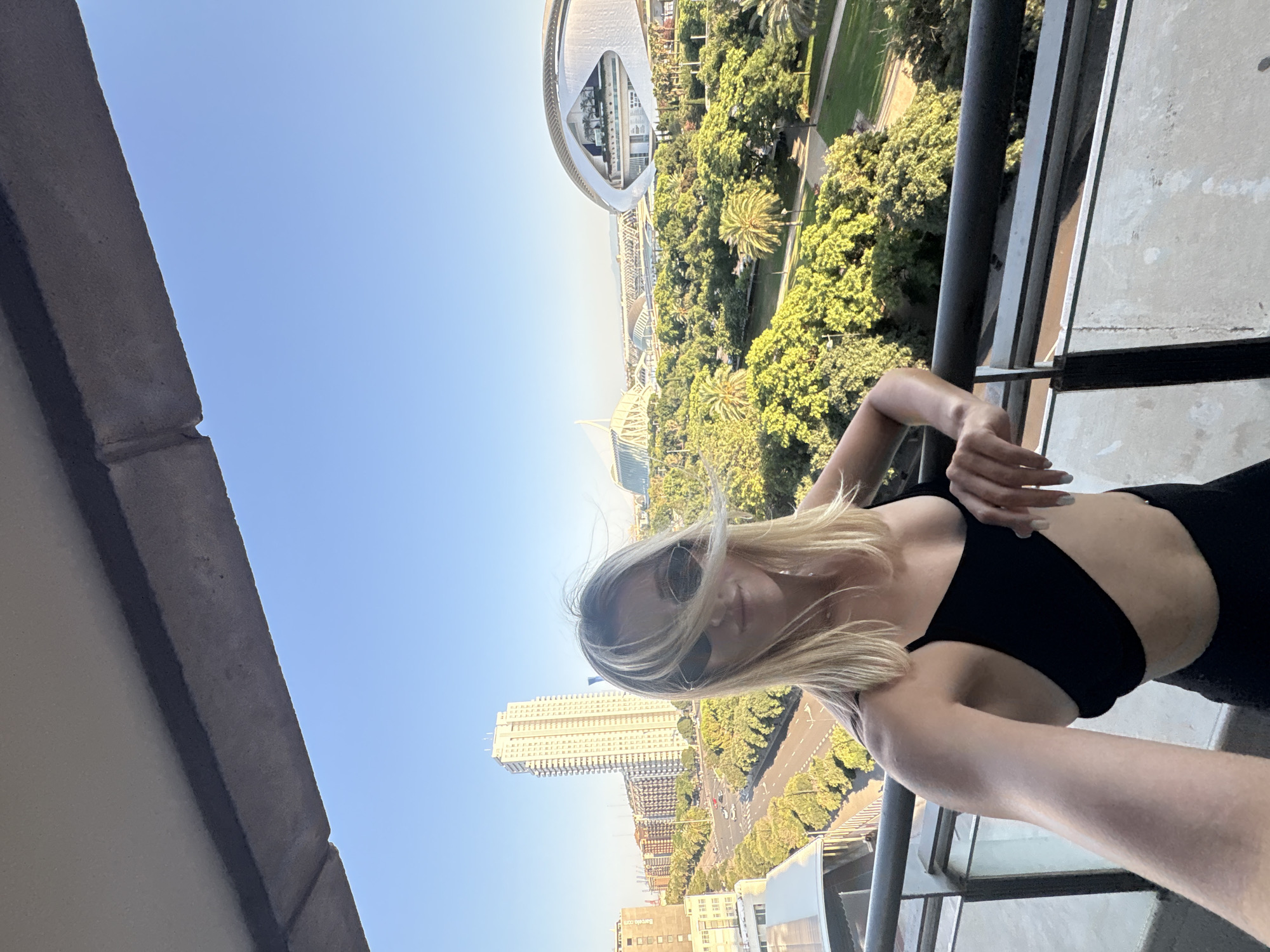 A blonde woman wearing sunglasses and a black bikini top stands on a balcony overlooking the City of Arts and Sciences in Valencia, Spain. The architecture of the City of Arts and Sciences, including the Hemisfèric and the Palau de les Arts Reina Sofía, is visible in the background, surrounded by lush green trees and a clear blue sky. A street with cars and palm trees runs parallel to the balcony. The woman is holding onto the railing with her left hand. The image appears to be a selfie, taken from a slightly low angle.
