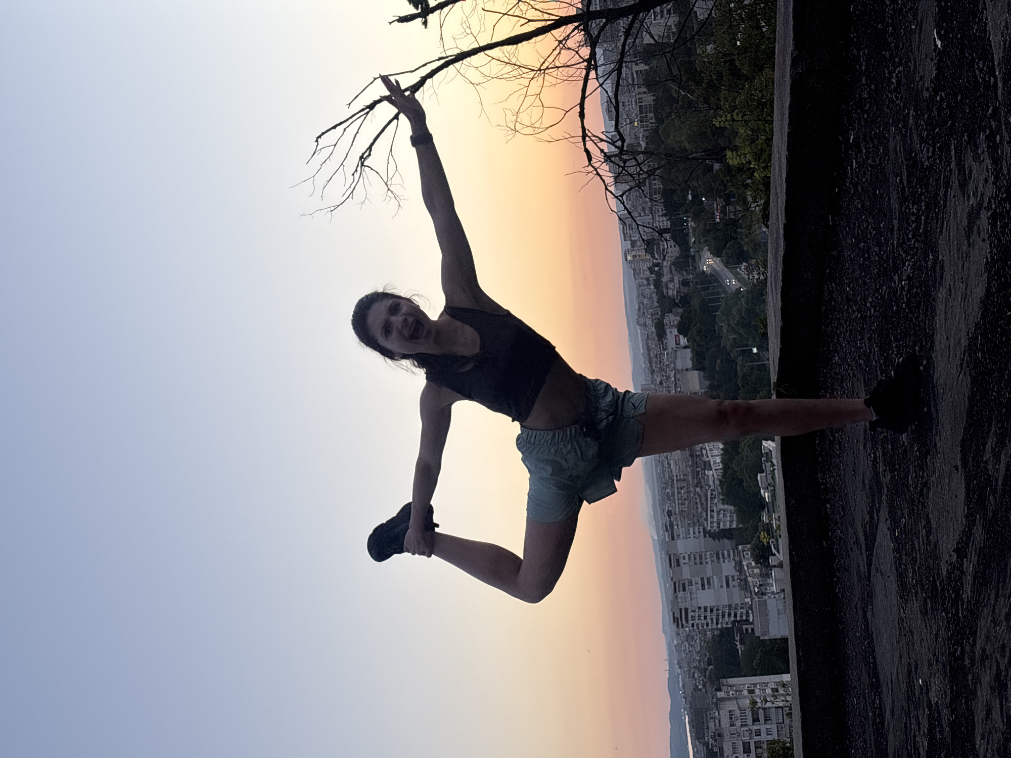 A woman is silhouetted against a sunset sky, performing a yoga pose on a rooftop overlooking the city of Varna, Bulgaria. She is wearing a dark tank top and light blue shorts, with her left leg bent and held behind her by her left hand, while her right arm is extended to the side. Her mouth is open in a shout or expression of exertion. The city below is a sprawl of buildings and trees, with a road visible in the middle ground. A bare tree branch extends into the frame on the right. The sky transitions from a pale blue at the top to shades of orange and pink near the horizon. The rooftop surface is dark and textured, appearing to be asphalt or concrete.