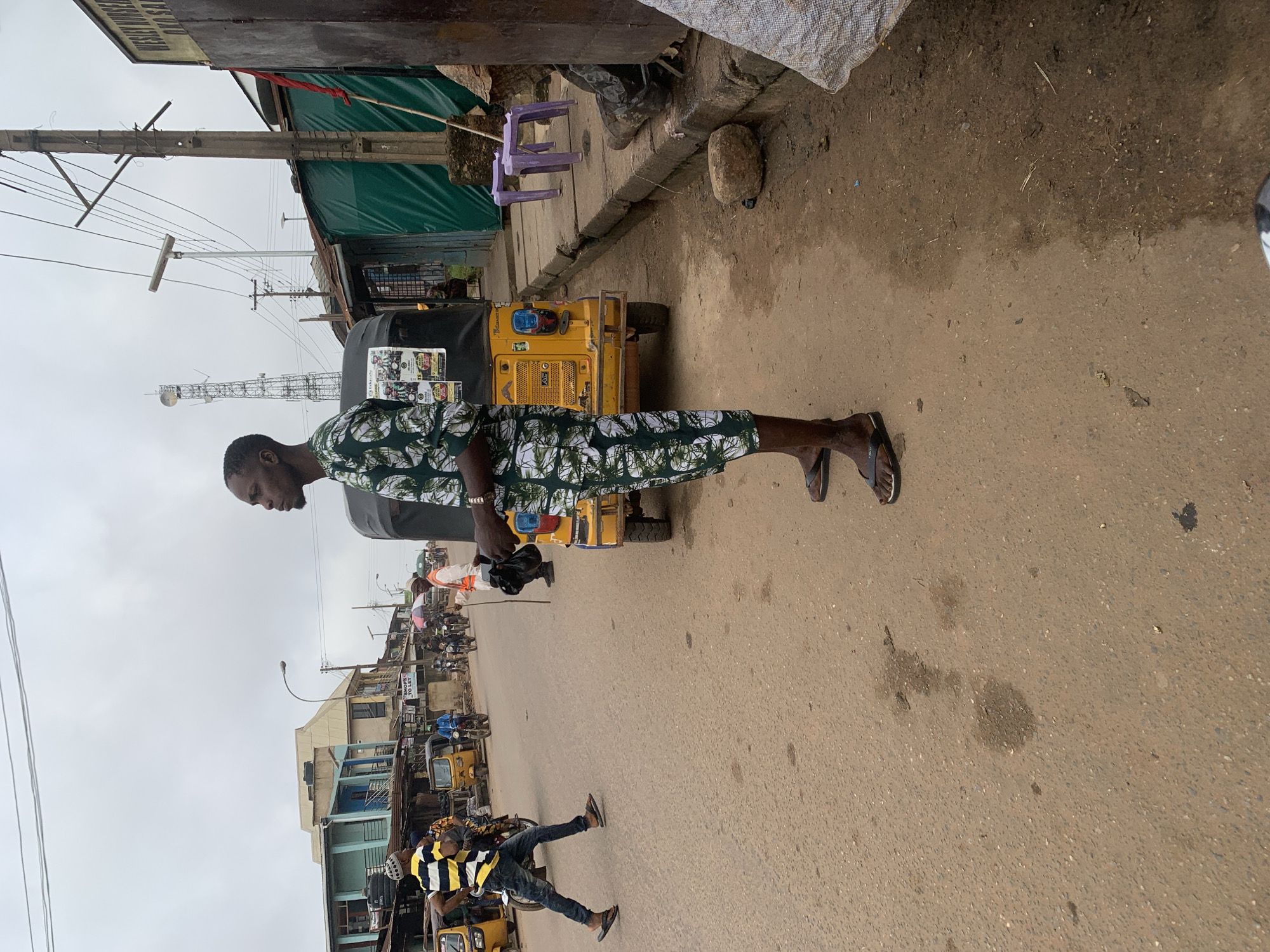 A full-shot, eye-level, outdoor photograph captures a man standing in profile on a dirt and asphalt road in Shagamu, Nigeria. The man, positioned in the right-center of the frame, is wearing a green and white patterned outfit that covers his arms and legs, along with black flip-flops. His left hand is relaxed at his side, while his right hand is partially visible near his waist. Behind him, to his left, a yellow motorized tricycle, known as an "okada" or "keke," is parked. The road is lined with various shops and buildings, including a green tarp-covered structure on the right and a light blue building with metal shutters on the left. Several other individuals and vehicles, including motorbikes and other tricycles, are visible in the background, suggesting a bustling commercial area. The sky is overcast with clouds. The ground is uneven, with patches of dirt, gravel, and some darker stains that appear to be wet spots or oil spills. Purple plastic stools are visible on the sidewalk to the right of the tricycle. The scene depicts a typical street view in a Nigerian city with active commerce and transportation.