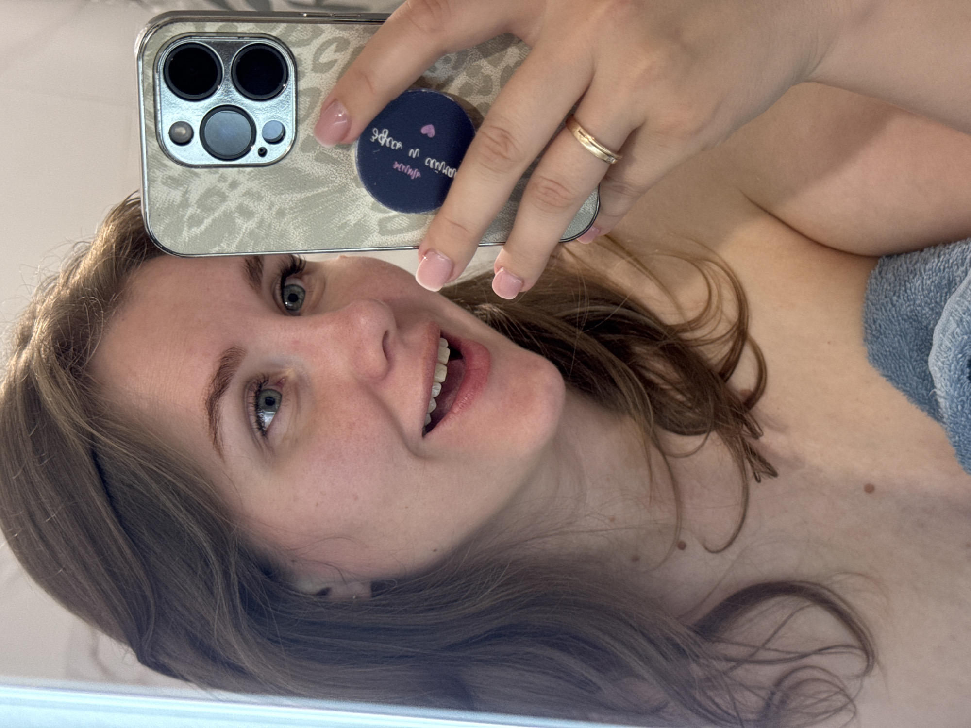 A woman is taking a selfie with a smartphone. The phone has a patterned case and a grip on the back. The woman has light skin, brown hair, and is wearing a gold ring. A blue towel is visible near her shoulder. The background appears to be a bathroom. There's no clear indication of the location being Ферма, Russia.