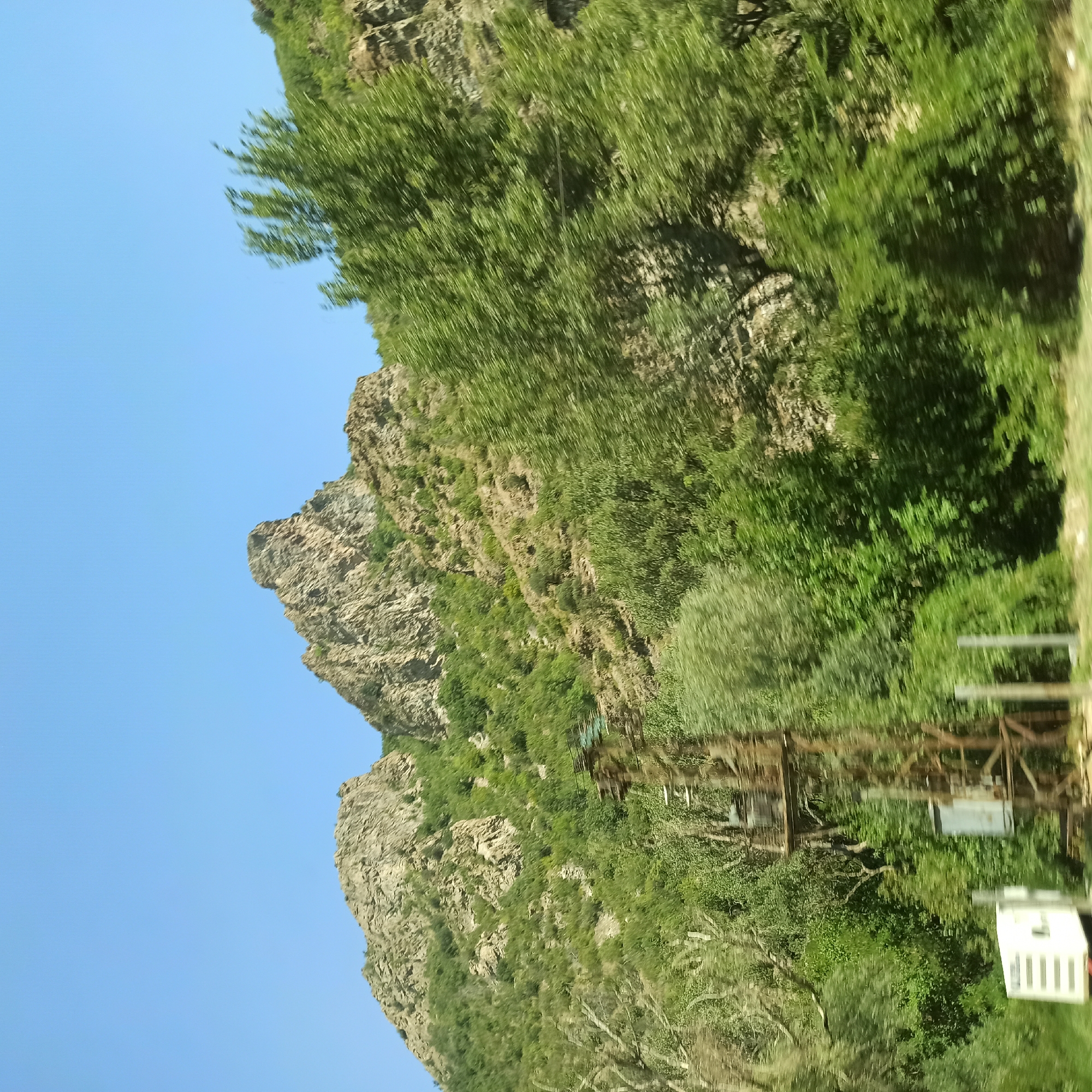 The image shows a natural landscape with a clear blue sky. In the foreground, on the left side, there is a white rectangular object, possibly a vehicle, with horizontal stripes. Next to it, there is a tall, rusty metal structure, possibly a power pylon, with some visible electrical equipment. The middle ground and background are dominated by a steep, rocky mountain covered with lush green vegetation. The trees appear to be in motion blur, suggesting the photo was taken from a moving vehicle. The location is identified as Krupnik, Bulgaria.