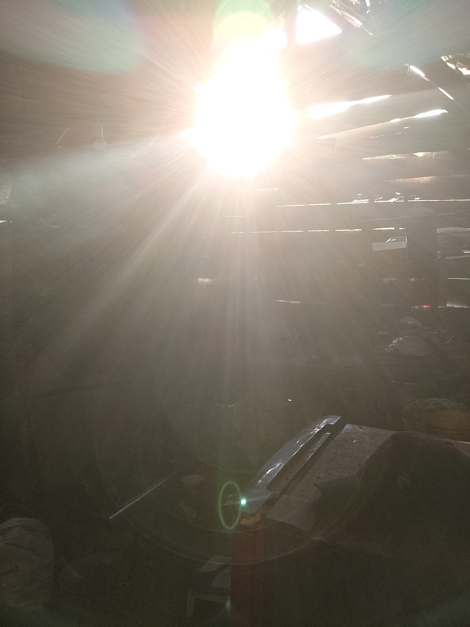 This image depicts a scene within a structure, likely a workshop or industrial space, illuminated by strong overhead sunlight streaming through gaps in the roof. The sun is the primary light source, creating bright flares and visible rays. In the foreground, a dark, irregular-shaped object is visible, possibly a workbench or a large piece of equipment, with a shiny metallic object resting on its surface. To the left of this, a large dark circular object is partially visible. The background is obscured by shadow and dust or smoke particles suspended in the air, giving the atmosphere a hazy, dramatic quality. The overall impression is one of a functional, somewhat cluttered, and dimly lit interior space, heavily affected by intense natural light. The location in Wadang, Indonesia, suggests a potentially tropical environment where such lighting conditions might be common.