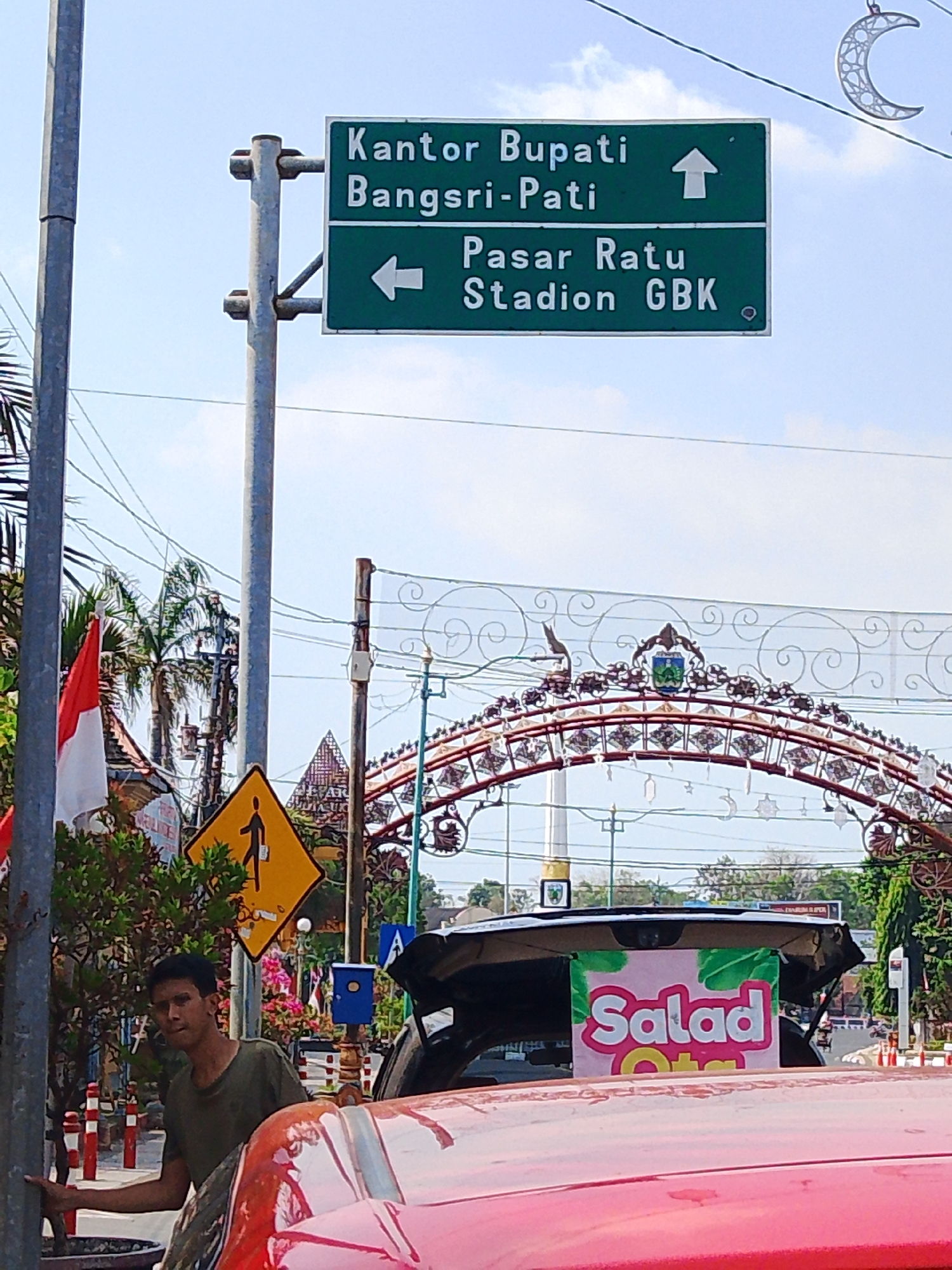 In Jepara, Indonesia, a street scene unfolds with a green directional signpost directing traffic. The upper panel indicates "Kantor Bupati" (Regent's Office) with an upward arrow, and the lower panel indicates "Pasar Ratu" (Ratu Market) and "Stadion GBK" (GBK Stadium) with a leftward arrow. Adjacent to the signpost, a red and white Indonesian flag flies. A yellow diamond-shaped pedestrian crossing sign is visible. In the foreground, a man in a green t-shirt stands near a red vehicle, with a sign reading "Salad Otak" on the back of a vehicle. An ornate archway with decorative elements and a coat of arms spans the background, suggesting a prominent location or event. The sky is partly cloudy, and power lines crisscross the scene.