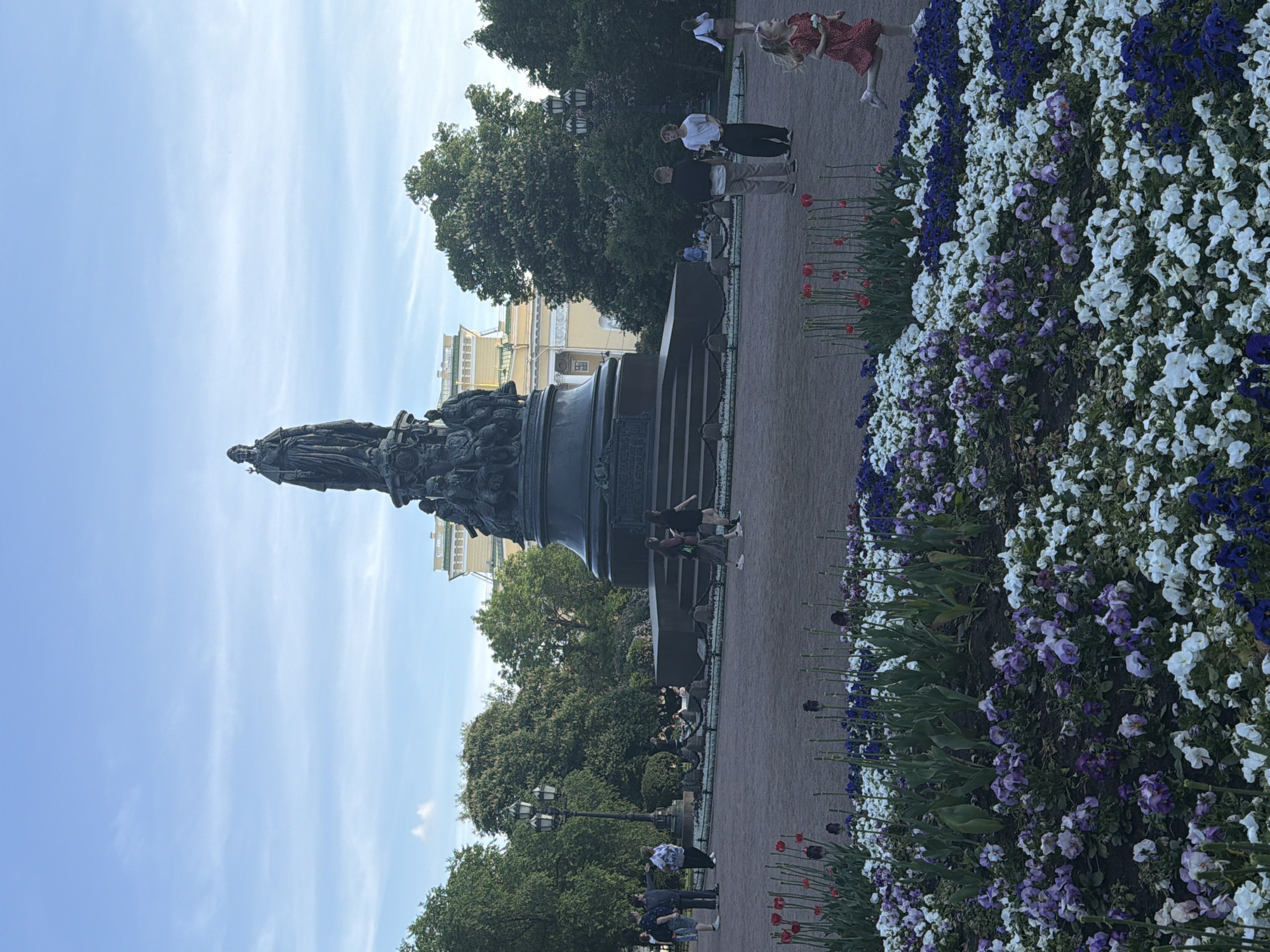 This image captures Ostrovsky Square in Saint Petersburg, Russia, centered on the Catherine the Great Monument. The sky is light blue with scattered wispy white clouds.
In the mid-ground, the large bronze Catherine the Great Monument stands prominently. The empress is depicted standing atop a dark, multi-tiered stone pedestal, holding a scepter. Bronze figures representing notable individuals from her reign are arranged around the pedestal's base. Behind the monument, a multi-story, light-colored classical building with a green roof is visible, likely the Russian National Library or Alexandrinsky Theatre.
The square's ground consists of a wide, light brown, packed earth or gravel surface. Numerous mature deciduous trees with green foliage densely surround the central area and monument. Several ornate black lampposts are positioned throughout the square.
In the foreground, an extensive flowerbed is visible. It features dense plantings of white, purple, and dark blue pansies or violas. Behind these, stalks of red tulips are present, some with existing blooms and others with dark, unopened buds.
Several individuals populate the scene. In the foreground-right, a young girl with light hair, wearing a red polka-dot dress, is running towards the right side of the frame, holding a small white object in her right hand. In the mid-ground-right, two men stand facing each other near the monument's base; one in a black shirt and light pants, the other in a white t-shirt and dark pants. On the mid-ground-left, a group of people is visible, including a woman in a light blue top and dark pants, and another woman in a dark top and striped skirt walking right. Additional people are scattered throughout the square, walking or standing.