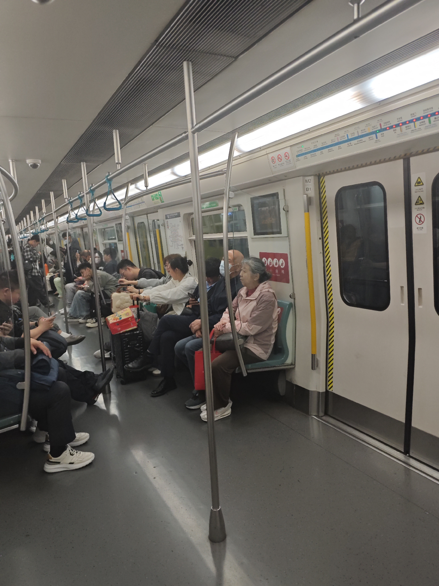 The image shows the interior of a subway car with passengers, likely in an urban setting. The subway car has seats along the side, and many passengers are seated or standing, occupying the available space. Some people are wearing masks. Handrails and poles are present for support, and there's a route map visible above the doors. Suitcases, bags, and various personal items are scattered around.