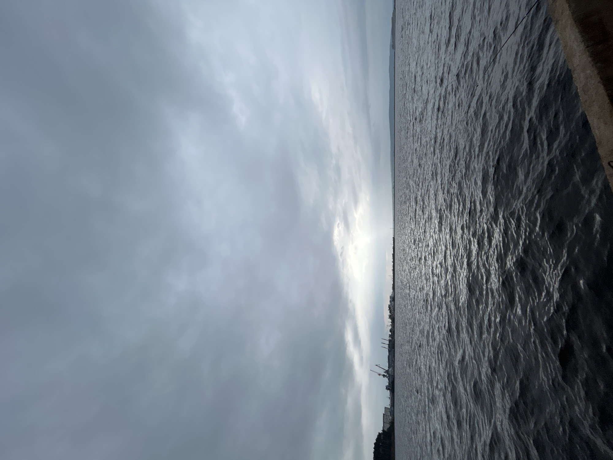 The image captures a wide view of a body of water under a cloudy sky. In the distance, along the horizon, a cityscape is visible, characterized by industrial structures such as cranes and buildings, suggesting a port or industrial area. The water surface is rippled, indicating moderate movement. The sky is overcast with a mix of dark and lighter clouds, with a bright spot suggesting a break in the clouds or the sun attempting to shine through. The overall atmosphere is somber due to the cloudy conditions. The foreground shows a concrete embankment, suggesting the vantage point is from a pier or waterfront structure. The scene is likely located in Chaikovsky, Russia, given the industrial elements and the vastness of the water body, which could be a reservoir or a large river. There are no people or specific actions clearly depicted.