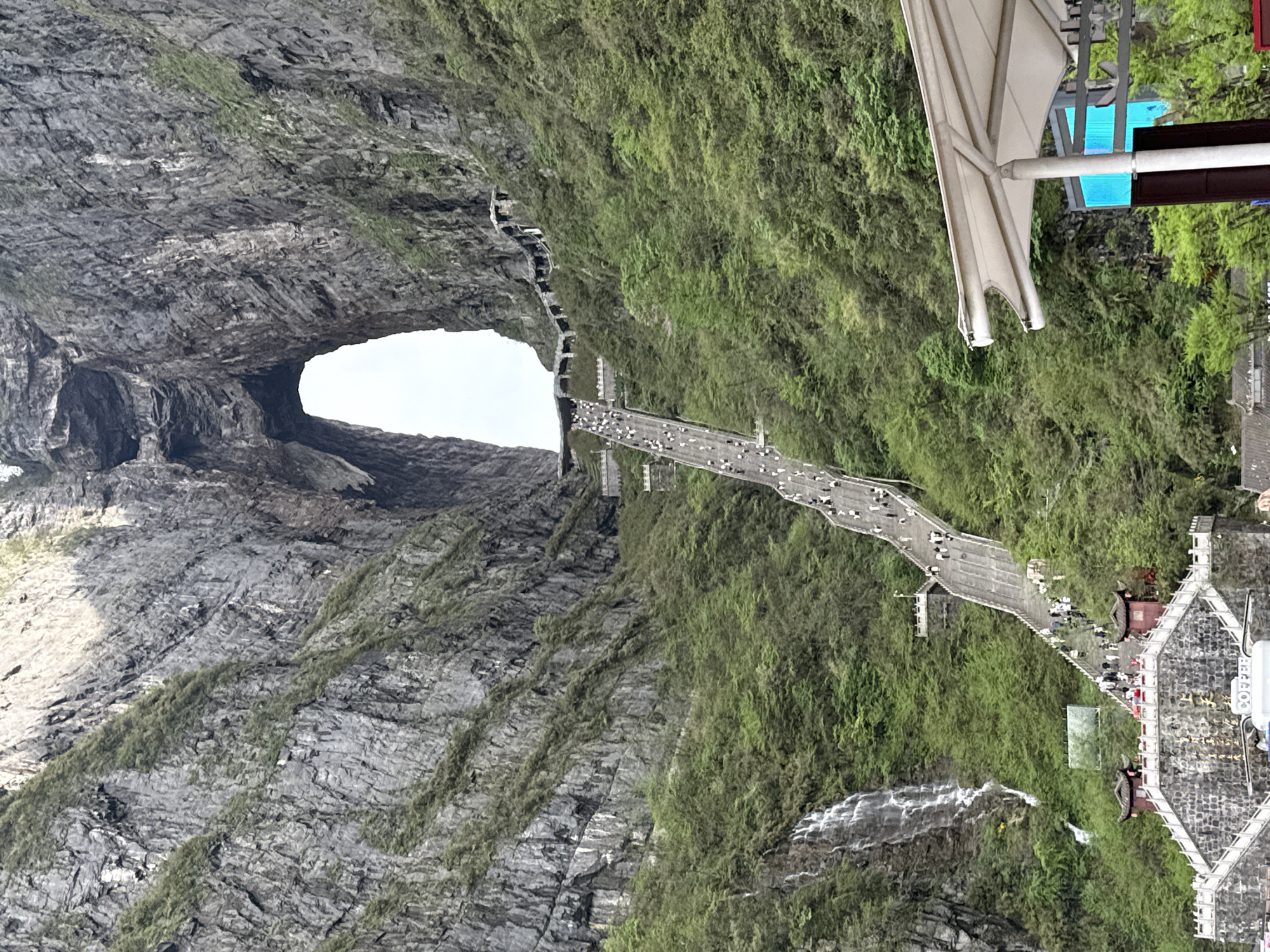 The image shows Tianmen Cave, also known as Heaven's Gate, a large natural arch in the Tianmen Mountain National Park in Zhangjiajie, Hunan Province, China. A pathway with numerous people walking on it leads towards the arch. Lush vegetation covers the cliffs alongside the pathway. Below the pathway, there is a waterfall and a traditional Chinese-style building. Part of a modern canopy structure is visible in the upper right of the image. The sky is white where it is visible through the arch. The cliffs surrounding the arch are predominantly gray with textured rock formations.