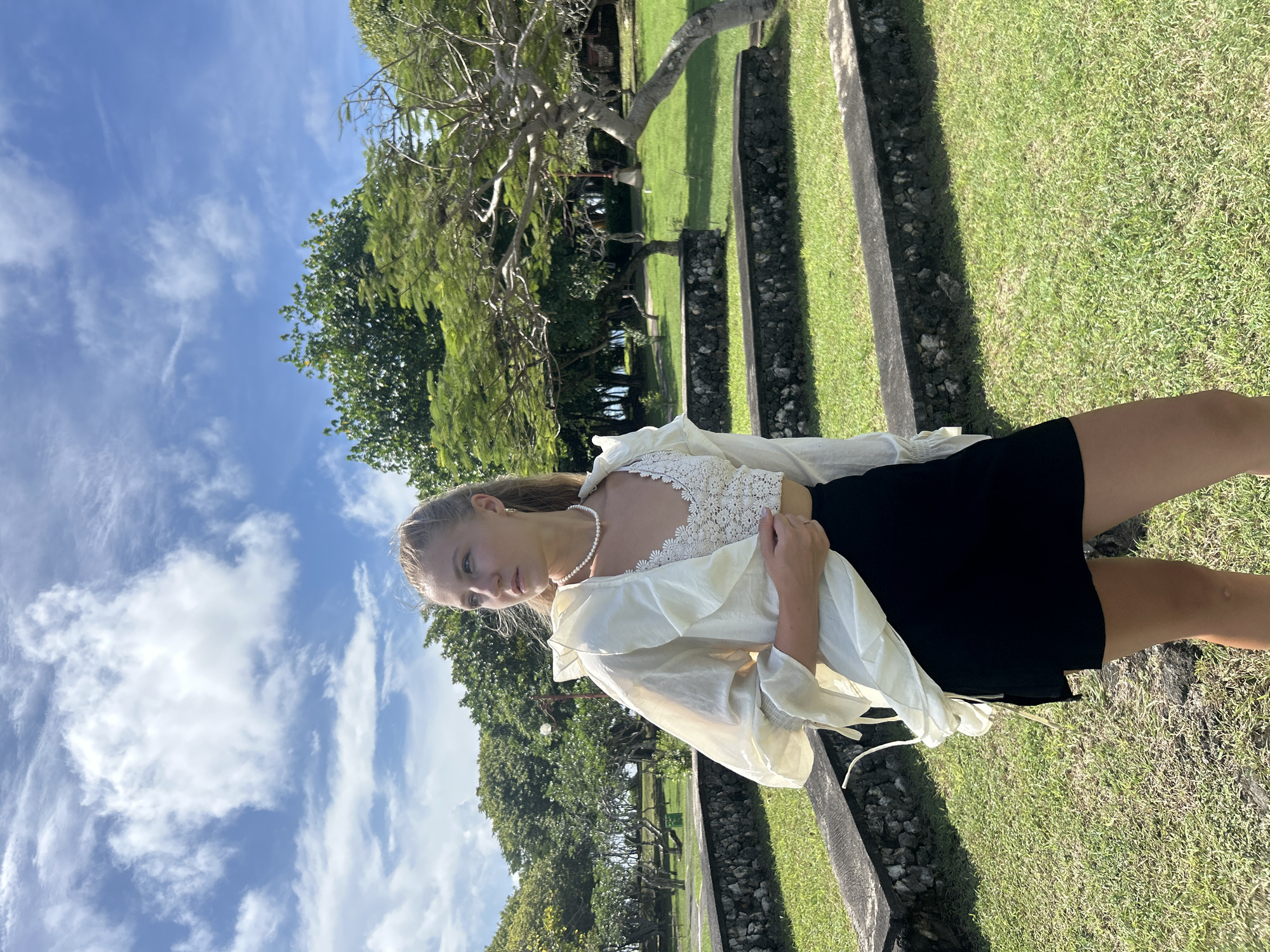 A photograph shows a light-skinned young woman standing on a grassy area in Kuta Selatan, Indonesia, under a partly cloudy sky. She is wearing a pearl necklace, a white lace top, a white sheer button-down shirt, and a black mini skirt. The grassy area is bordered by stone features. Trees are visible in the background.