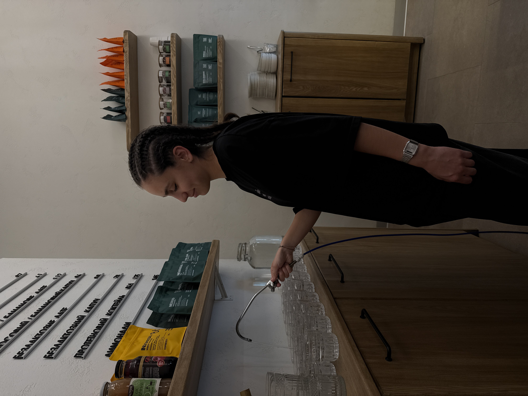 The image displays the interior of a commercial establishment, specifically a cafe or health-focused beverage bar, located in Uzhhorod, Ukraine. The scene features a young adult female, depicted in profile facing right, actively filling a glass with water. She has dark hair styled in braids and is wearing a black short-sleeved top with a silver-toned, rectangular digital watch on her left wrist. Her right hand holds a flexible, chrome-plated faucet, from which a stream of water descends into a clear, textured glass positioned on a light wooden counter.
The establishment's decor includes textured white walls and light-colored wooden shelving and cabinetry. A wall-mounted menu board on the left displays items in Ukrainian with accompanying numbers, likely prices. Visible entries include "ОСОВИЙ / БАНАНОВЕ ЛАТЕ" (Oat / Banana Latte), "БЕЗЛАКТОЗНЕ ЛАТЕ" (Lactose-Free Latte), "БЕЗЛАКТОЗНИЙ КАПУЧ" (Lactose-Free Cappuccino), "ПРОТЕЇНОВИЙ КОКТЕЙЛЬ" (Protein Shake), and "АММОНАЙ".
Below the menu, a wooden shelf holds various retail products: two glass bottles, one prominently labeled "СІК ГРУШЕВИЙ" (Pear Juice), and multiple pouches of coffee beans. Labels visible on these coffee pouches include "INDONESIA", "MEXICO", and "NICARAGUA", often alongside the term "ESPRESSO".
On the wooden counter, a row of clear, identical textured glasses is neatly arranged. Behind the faucet, a large clear glass jar with a lid is present. A blue hose connects to the base of the faucet and extends downwards behind the counter.
To the right of the central figure, two additional wooden shelves are affixed to the wall. The top shelf holds rows of small pouches, some dark green and some orange. The lower shelf contains several small, clear jars with labels, and more dark green coffee pouches, with "COLOMBIA" and "INDIA" visible on some. Below these shelves stands a light-colored wooden cabinet with black pull handles. On top of this cabinet, a stack of white plates, several white bowls, and a glass containing utensils are organized. The floor is light-colored and appears to be tiled.