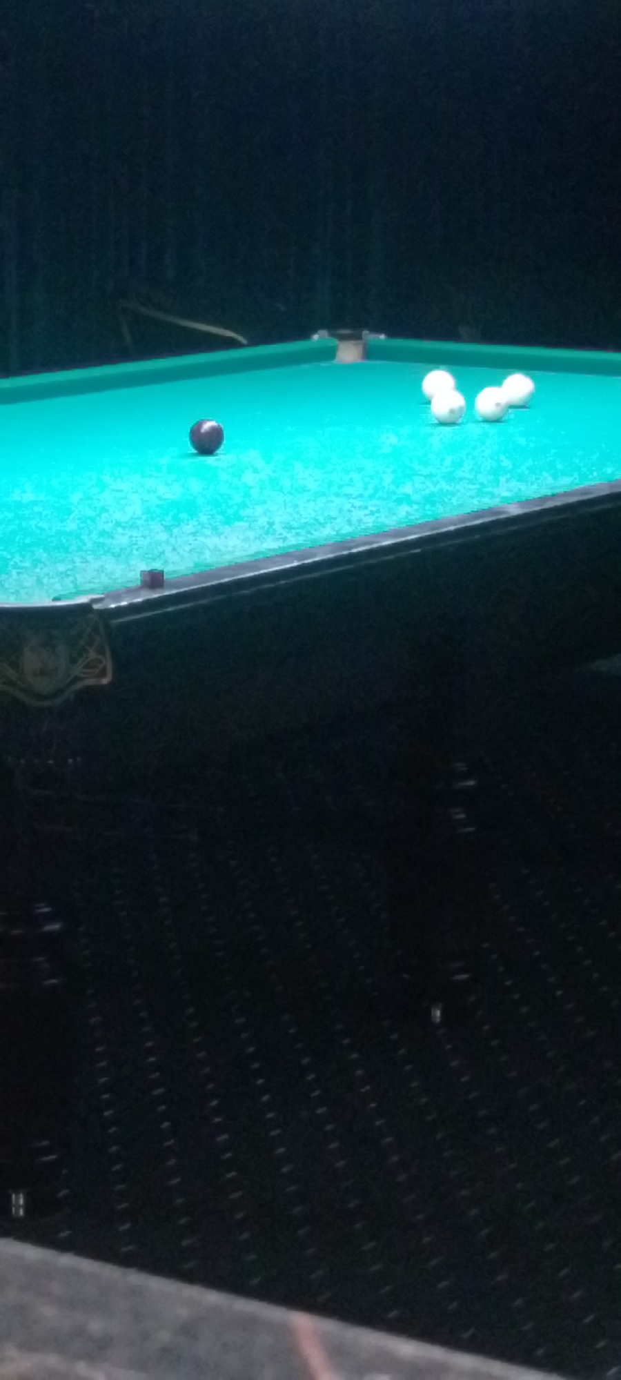 The image shows a billiard table with a green felt surface. Several white billiard balls are scattered on the table, along with a single dark-colored ball positioned near the center. The background is dark and appears to be a dimly lit room, possibly a bar or game room. The lighting suggests it is evening or nighttime. The provided metadata indicates this image was taken in Angren, Uzbekistan.