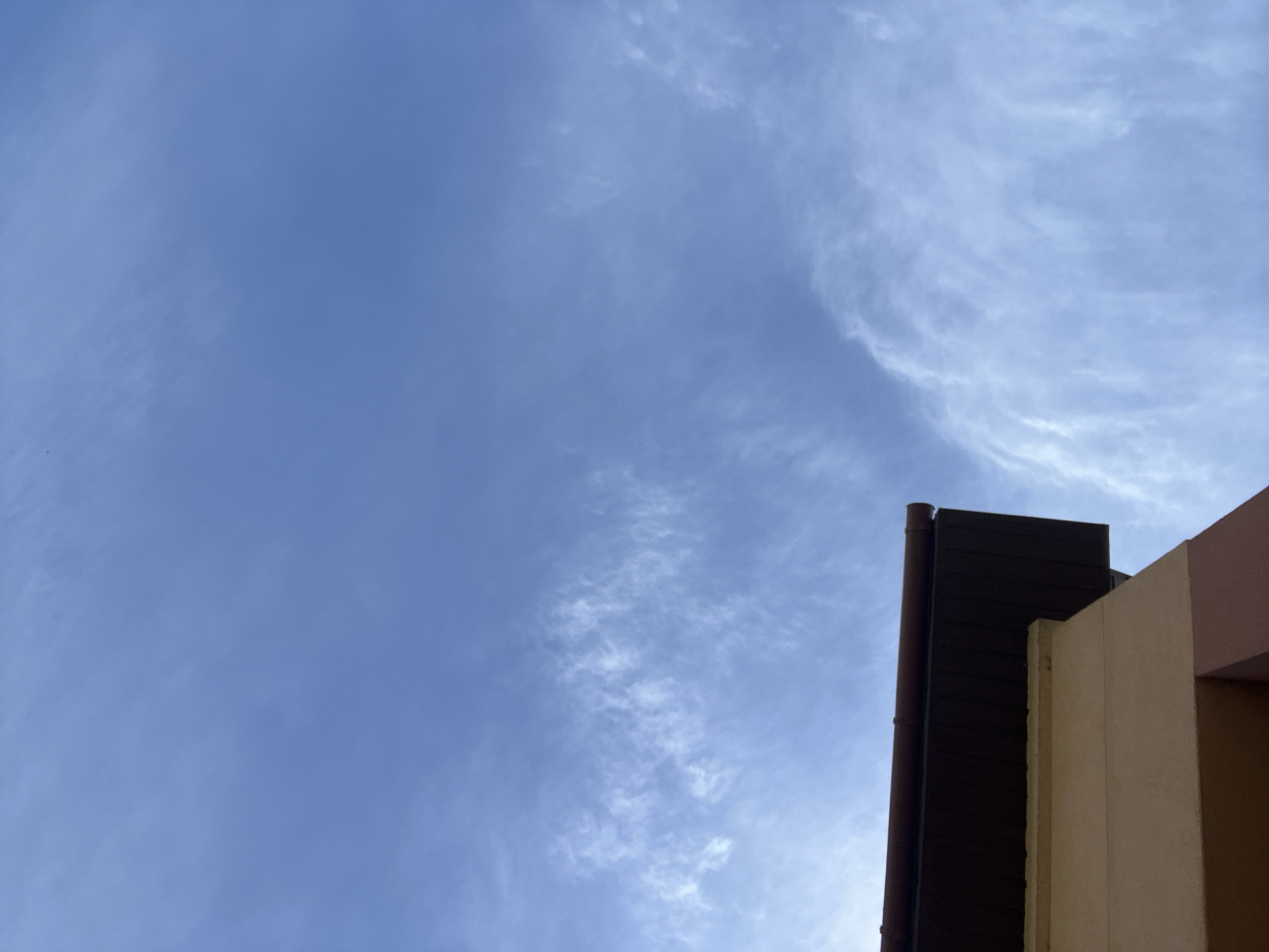 The image shows a view of the sky in Onokivtsi, Ukraine. The sky is blue with thin, wispy clouds. In the lower right corner of the image, a portion of a building is visible. It appears to be a multi-story structure with different colored sections - brown, tan, and pink. A drainpipe is also visible alongside the building's brown section.