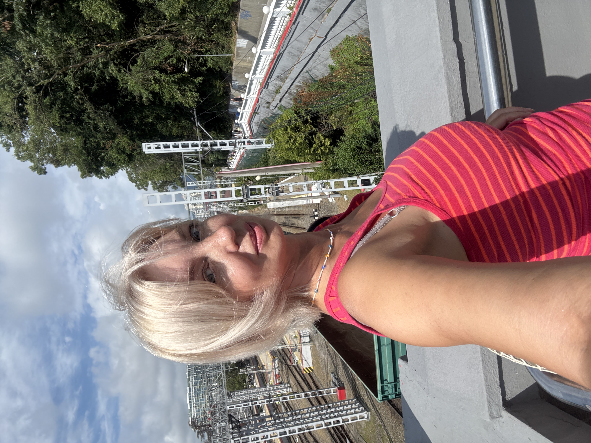 A blonde woman with fair skin and freckles is taking a selfie outdoors. She is wearing a pink and orange striped tank top and a beaded necklace. Behind her, there is a train station with tracks, overhead wires, and buildings in the distance. A lush green hillside with trees is visible to the right, and a white railing with red trim lines a pathway. The sky is partly cloudy.