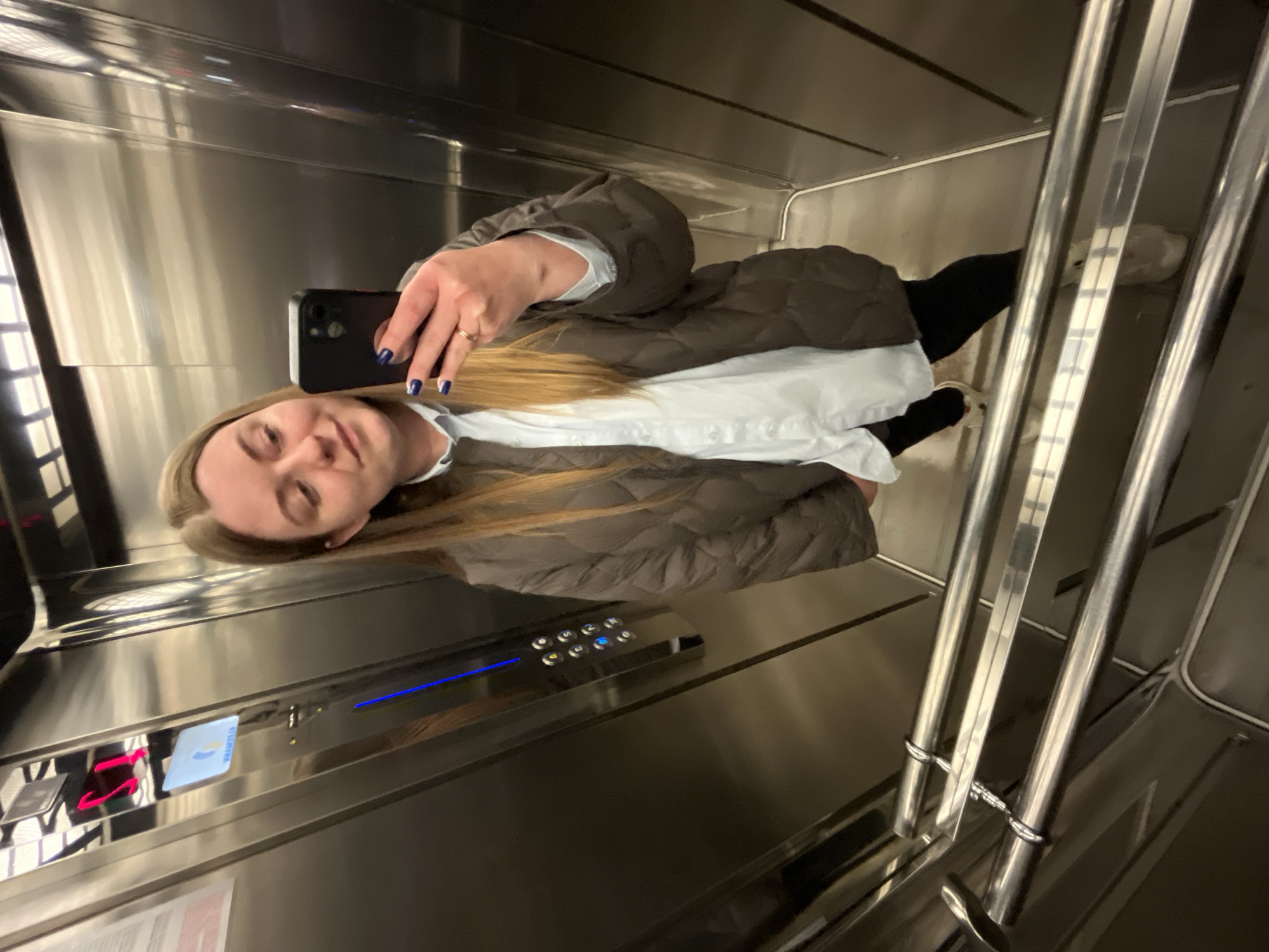 A person takes a mirrored selfie inside an elevator. The subject is fair-skinned with blonde hair and wears a brown puffer jacket over a white shirt and black pants. They are holding a black smartphone. The elevator interior is stainless steel, with visible control panels. The location might be inferred as Klin, Russia, based on context not explicitly visible in the image.