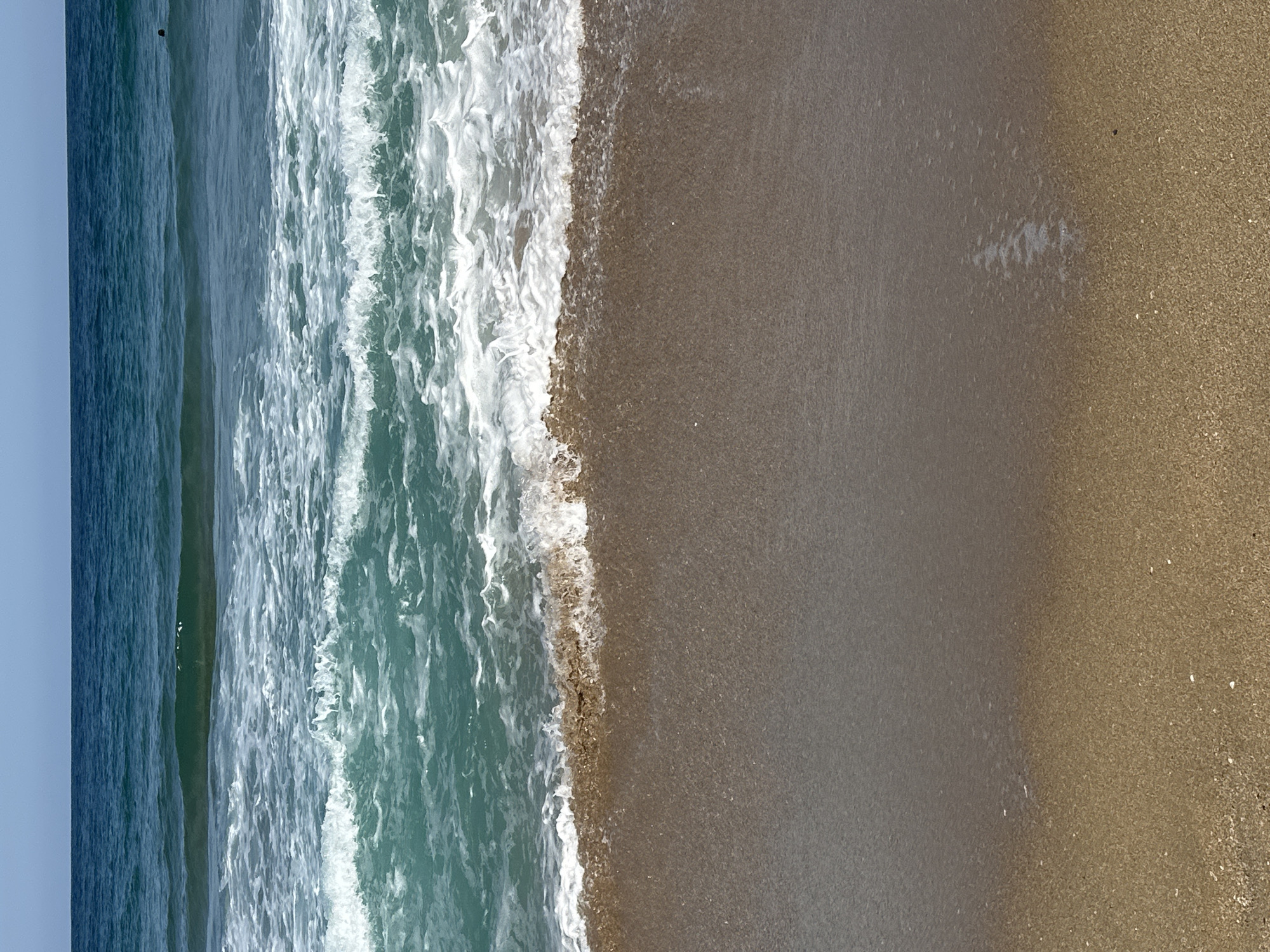 The image shows a view of the ocean from a beach. The foreground is a sandy beach with some small shells. A gentle wave is washing onto the shore, creating a line of white foam. The ocean water is a clear blue-green, with larger waves in the background. The sky above is a clear, pale blue. There is a person visible in the ocean in the distance, to the right of the frame, appearing as a dark silhouette.