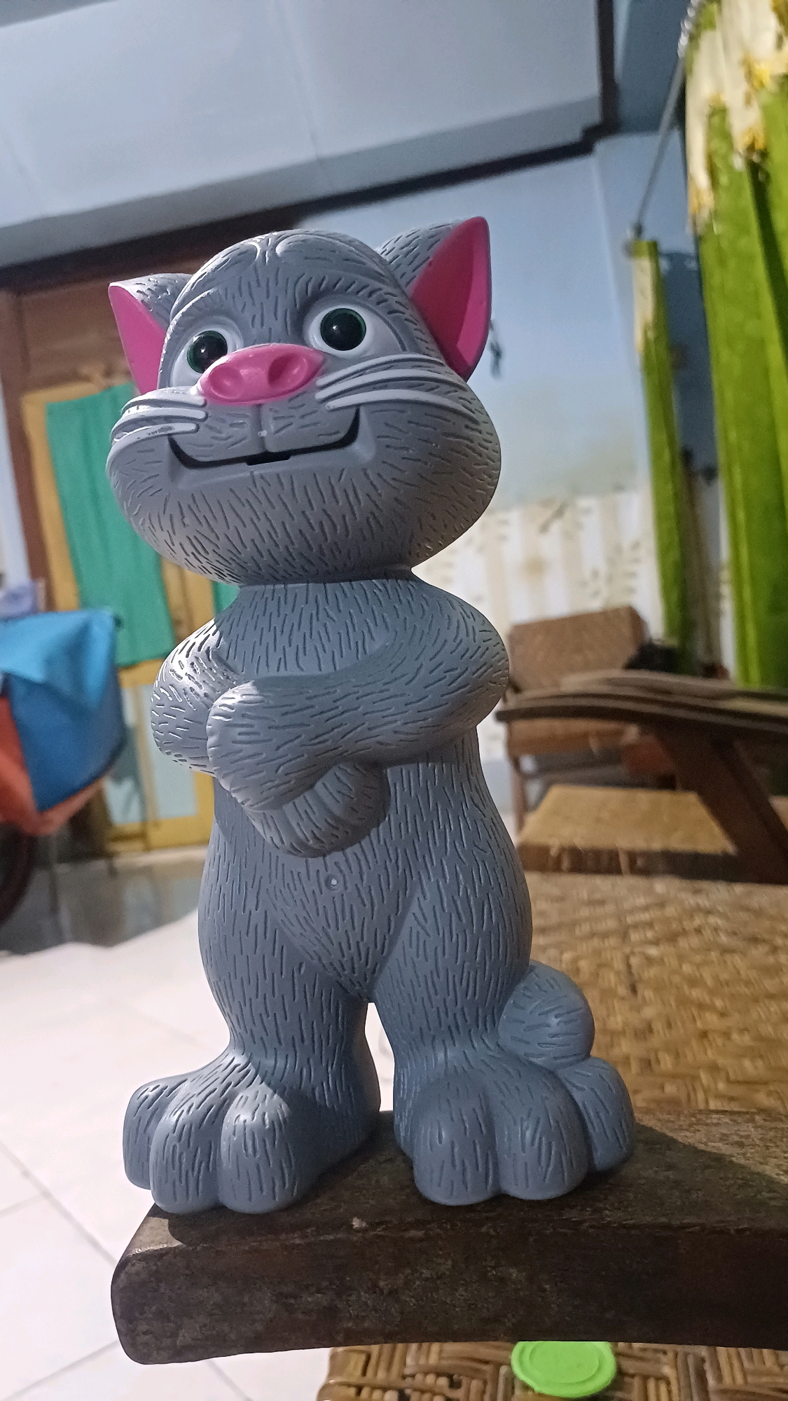 A gray cat figurine, resembling the character Tom from Talking Tom and Friends, stands on a dark wooden surface. The figurine has large green eyes, a pink nose, and pink inner ears. Its arms are crossed over its chest, and its body is textured to resemble fur. The background appears to be an indoor setting with green curtains visible. The lighting suggests it might be evening or a dimly lit room. The flooring visible in the lower left appears to be tiled.