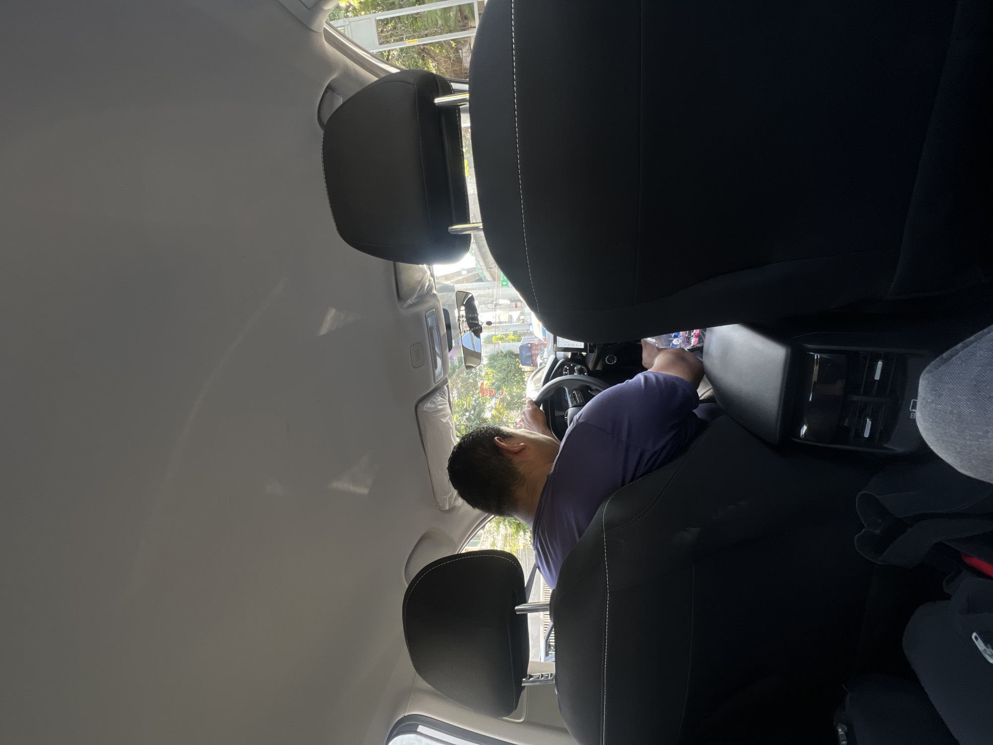 The image captures an interior view from the rear passenger seat of a car, looking towards the front. The primary subject is a person in the driver's seat, seen from behind. This individual has short, dark hair and is wearing a short-sleeved, light purple or blue shirt. Their hands are on the steering wheel, and they appear to be driving or waiting in traffic.
The car's interior is predominantly light gray for the ceiling and darker gray/black for the seats and dashboard. Two black headrests are prominent in the foreground, belonging to the front seats, with one closer to the camera. The driver's headrest is visible in front of the viewer. Through the windshield, an outdoor scene is visible, showing bright sunlight, green trees, and glimpses of buildings or structures, consistent with an urban or suburban environment like Mexico City.
Notable details include a rearview mirror with a hanging ornament, possibly a rosary, visible in the center of the windshield. Below the rearview mirror, parts of the dashboard and console are visible, including what appear to be air vents and possibly some bottles or containers in the center console or door pocket. To the right, on the passenger side floor, a black bag or backpack is partially visible. The overall lighting suggests it is daytime with bright sunshine. No discernible text is visible within the image.