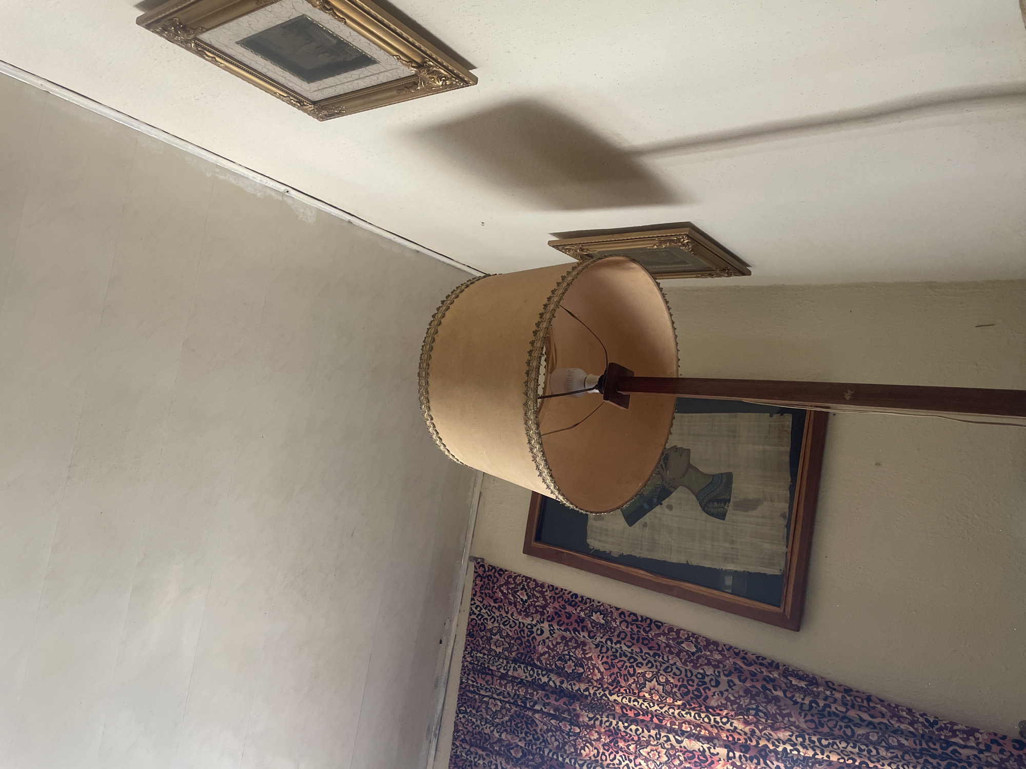 The image shows the interior of a room with cream-colored walls and a white ceiling. There are three framed pictures attached to the ceiling. A floor lamp with a tan shade is positioned in the center of the image. Beneath the lamp, a framed artwork of a woman's profile hangs on the wall. Leopard print curtains cover the lower section of the wall. The location is Gura Top, Nigeria. There is no evidence of any people in the image.