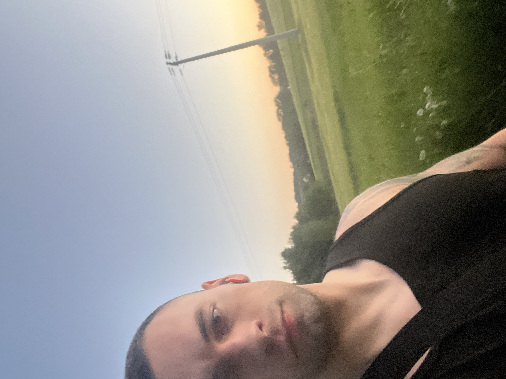A man wearing a black tank top is looking at the camera. He has short dark hair and stubble. The background shows a field, trees, and a utility pole against a sunset sky. The location appears to be a rural area. The city is Unknown, Unknown.