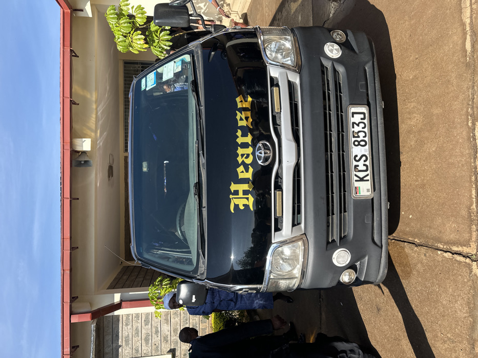 The image shows a black van with the word "Hearse" in gold letters on the front. The van is parked in front of a building. The van has a Kenyan license plate that reads "KCS 853J". The image is taken in front of a house in Githurai, Kenya, with coordinates latitude: -1.179, longitude: 36.914. The scene shows a person in a blue dress and a white head wrap standing by the van, while a person in a black suit stands in front of the van. The van is parked on concrete pavement in front of a building with a stone wall. The building has a red roof and is located on a residential street. The sky is blue and clear. There is a tree in the background behind the van.