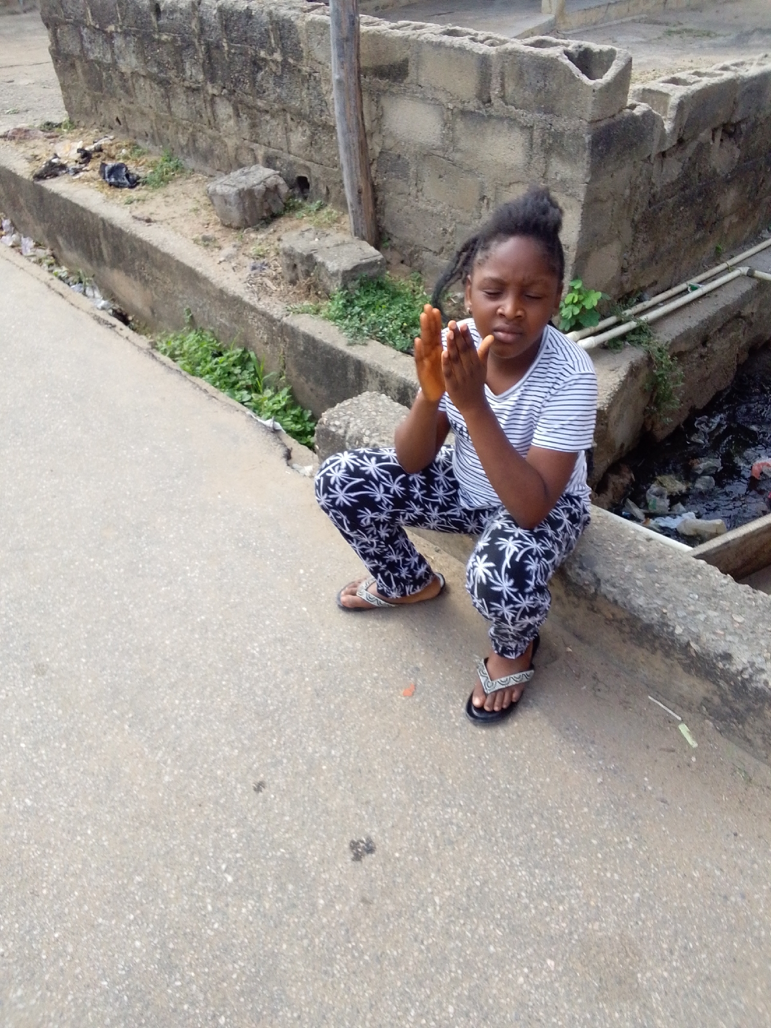 A young girl is captured crouching on a concrete ledge next to a drainage ditch in what appears to be an outdoor, semi-urban setting in Lokoja, Nigeria. She wears a white and blue striped short-sleeved t-shirt, black pants with white palm tree or star-like patterns, and mismatched sandals – one brown, one black and white. Her dark hair is pulled up, and several strands frame her face. Her eyes are closed, and her hands are clasped together in front of her chest, suggesting a moment of prayer or deep contemplation.
The immediate surroundings feature a rough concrete surface in the foreground, transitioning to a raised concrete ledge where the girl is positioned. To her right is an open drainage channel containing murky water, visible debris, and some green vegetation. Behind her, a wall constructed from grey concrete blocks shows signs of wear and appears somewhat unfinished, with some blocks exhibiting hollow centers. Sparse green plants grow in the crevices of the concrete and dirt. A wooden pole stands vertically against the background wall. The scene is brightly lit, indicating it is daytime with clear or lightly overcast weather. No visible text is present in the image.