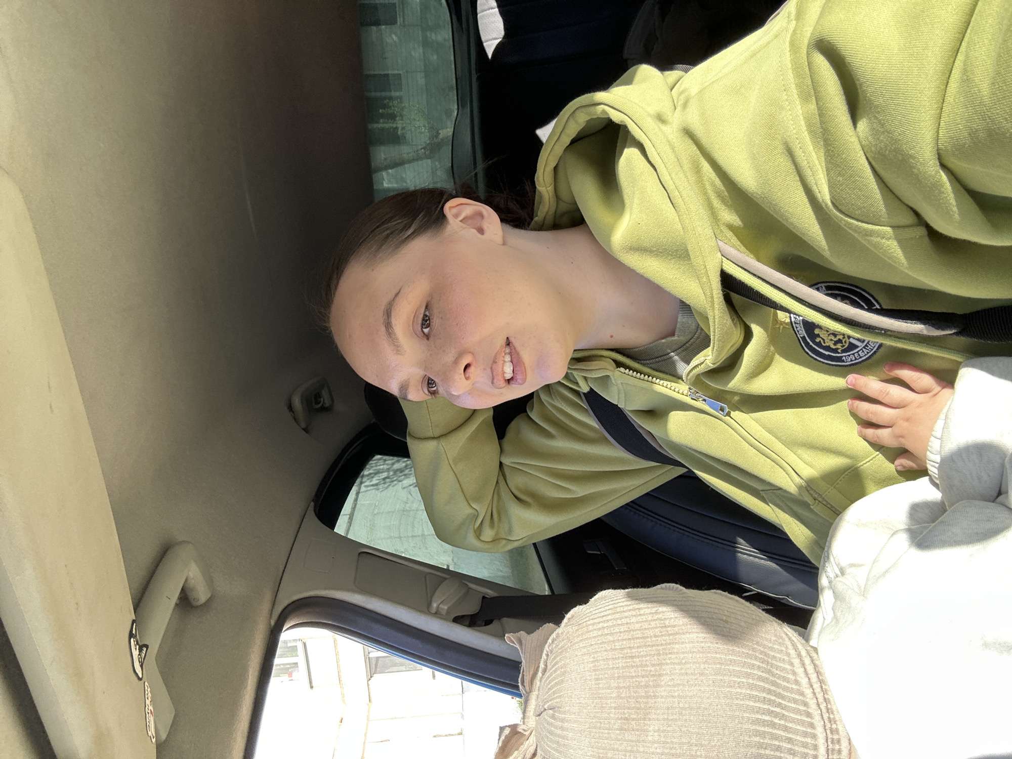 The image depicts a young woman inside a car. She is wearing a light green hooded sweatshirt. A small child's hand is visible on the woman's lap. The car's interior is visible, including the seat, window, and ceiling. Outside the window, buildings are visible, potentially within the city of Chaikovsky, Russia.