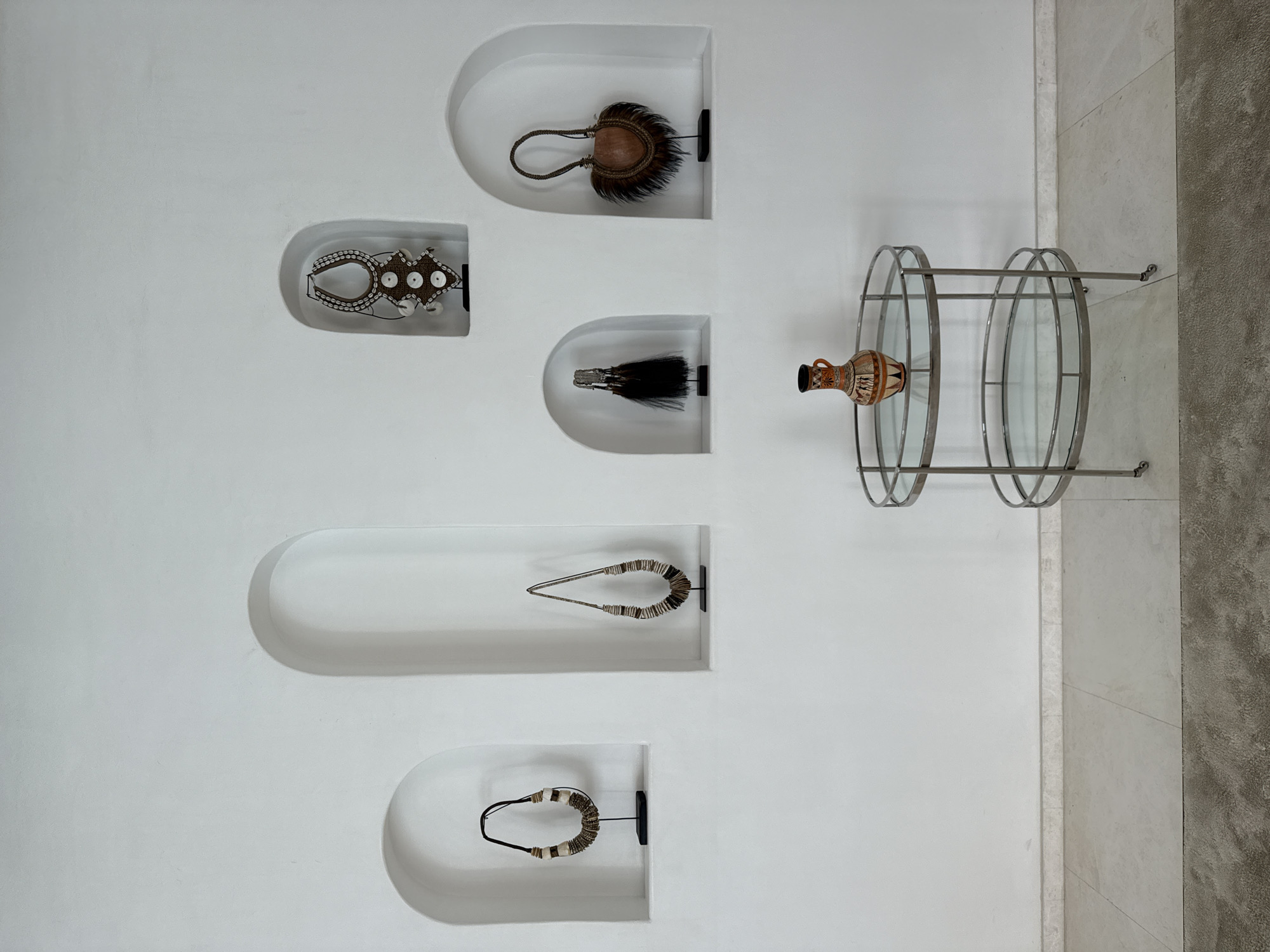The image shows a display of artifacts on a white wall, possibly in Ghadeer Al Tayr, United Arab Emirates. Six recessed niches contain various objects resembling tribal or ethnographic jewelry and adornments. The items include beaded necklaces, feathered objects, and possibly ceremonial ornaments. To the right of the niches is a metal and glass circular shelving unit holding a decorative vase or urn. The overall aesthetic is modern and minimalist, highlighting the artifacts as cultural or artistic objects. The wall is part of an interior space, with a section of tiled or stone wall visible to the right of the shelving unit.
