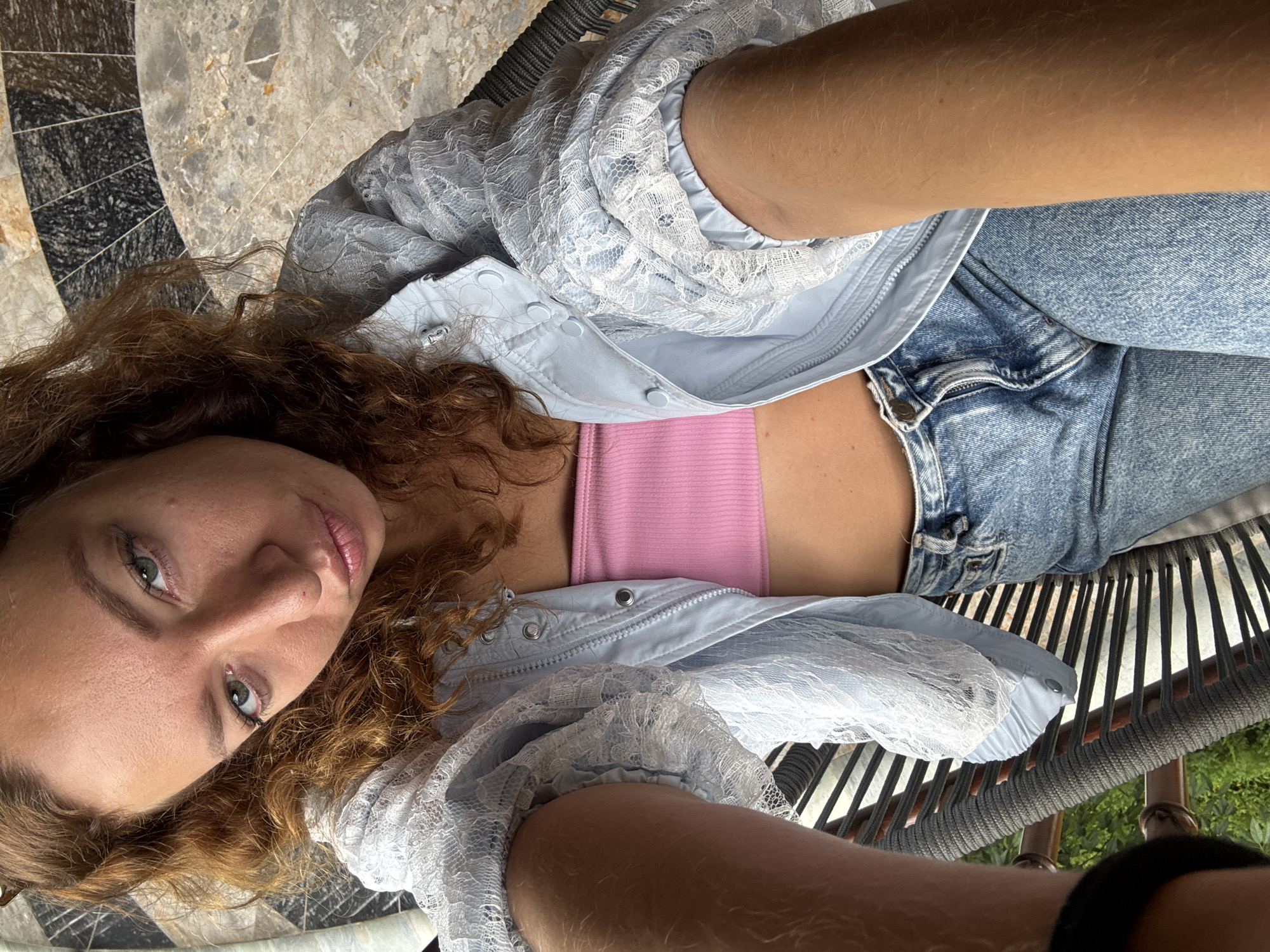 A young adult female is depicted taking a selfie, looking directly into the camera with her head slightly tilted to the left and lips subtly pursed, exhibiting a serious expression. She has long, wavy to curly, reddish-brown hair, parted on the left side. Her eyes are light-colored, appearing greenish-blue. Her skin is fair, with visible minor blemishes or freckles on her face.
She is wearing a pink strapless bandeau-style crop top, which is partially covered by a light blue bomber-style jacket. The jacket features a zippered front closure and snap buttons on the collar and cuffs. Its sleeves and portions of the back are made of a translucent white lace material. She is also wearing light blue denim jeans with a visible button and a partially unzipped fly. One arm, appearing tan and lightly haired, extends into the foreground from the lower left, holding the camera device out of frame.
She is seated in an outdoor chair with a dark gray woven or corded backrest and a partial armrest visible on the left. The background features a distinctive tiled floor with a geometric pattern composed of various shades of gray, black, and beige stone or marble, including curved and radial elements. Beyond the chair and to the left, green foliage from plants or trees is visible, indicating an outdoor setting. The scene is well-lit by natural light. The specific geographic location of Púng Luông, Vietnam, cannot be verified from the image content.