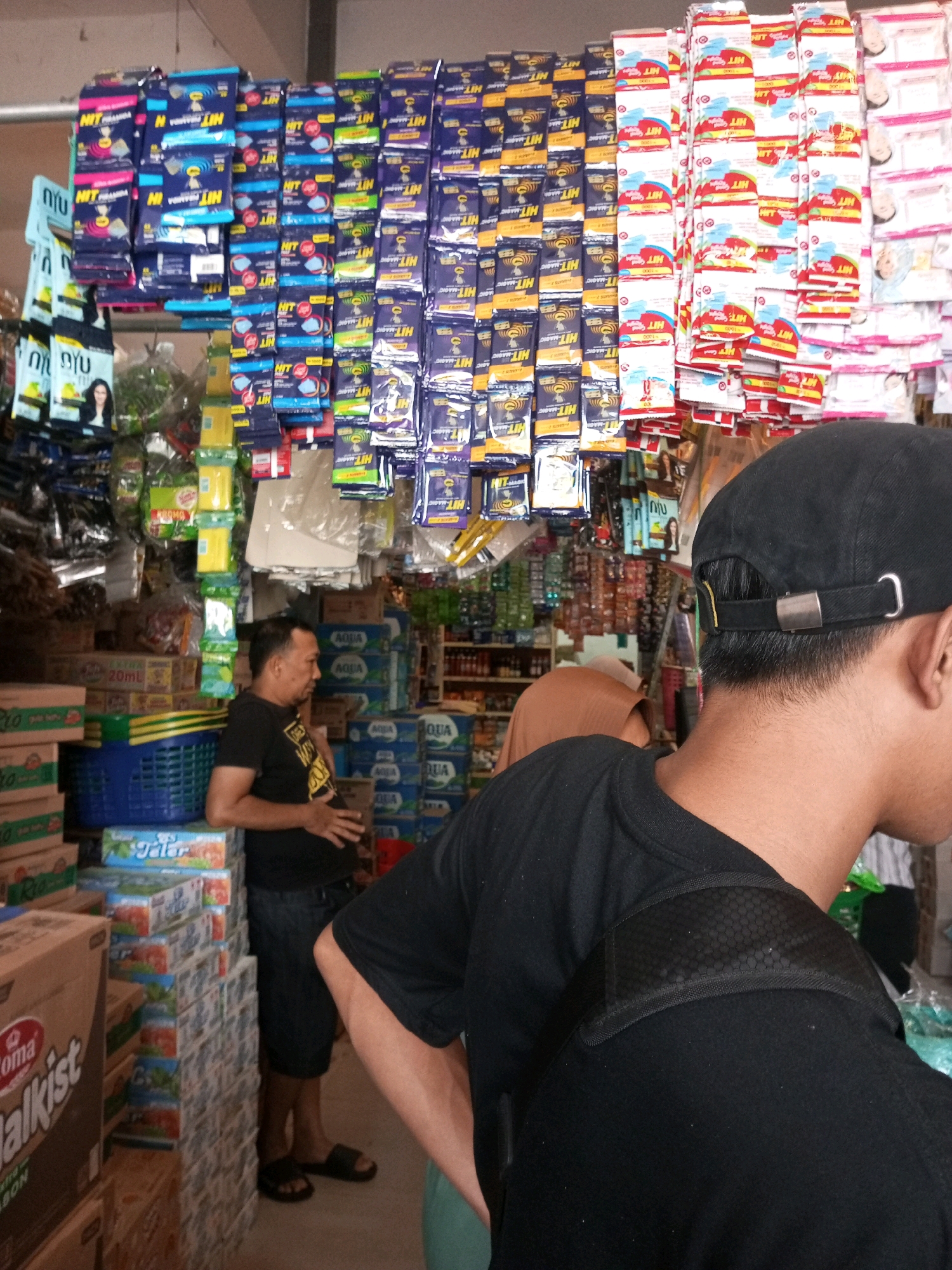 The image depicts the interior of a small retail store, likely a minimarket or warung, in Kepanjen, Indonesia. The shelves are stocked with a variety of packaged goods, including what appear to be household cleaning products, food items, and drinks. Prominently displayed are rows of small sachets of "HIT" brand mosquito repellent, in both blue and red packaging. Stacks of bottled water, possibly Aqua brand, are visible in the background. A man in a black t-shirt and dark shorts stands in the foreground, looking towards the right of the frame with his hands clasped. Another person, partially obscured, wearing a brown hijab and a light-colored garment, is seen in the middle ground to the right. The overall atmosphere suggests a typical scene of commerce in a local Indonesian community.