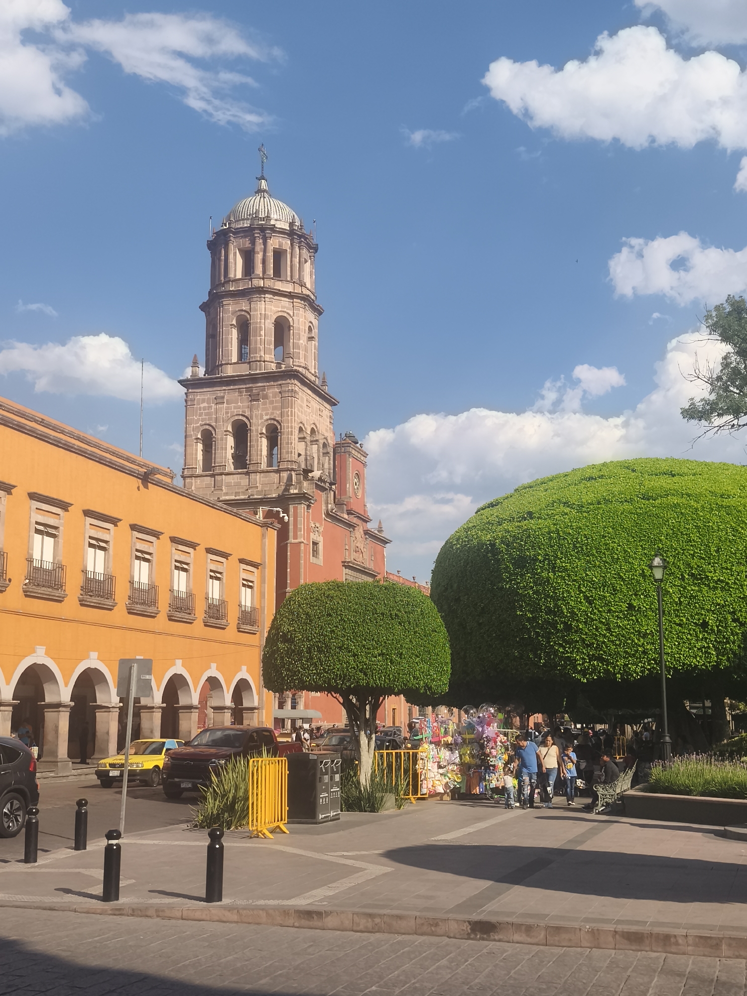 This image captures a sunny day in Querétaro, Mexico, featuring a prominent church tower and colonial architecture. The foreground shows a pedestrian area with large, meticulously trimmed trees, and vendors with their wares. People, including families, are walking and browsing. Behind them, vehicles, including a taxi, are parked along the street. The architecture consists of colorful buildings, primarily in shades of orange and red, with arches and windows. The sky is bright blue with scattered clouds.