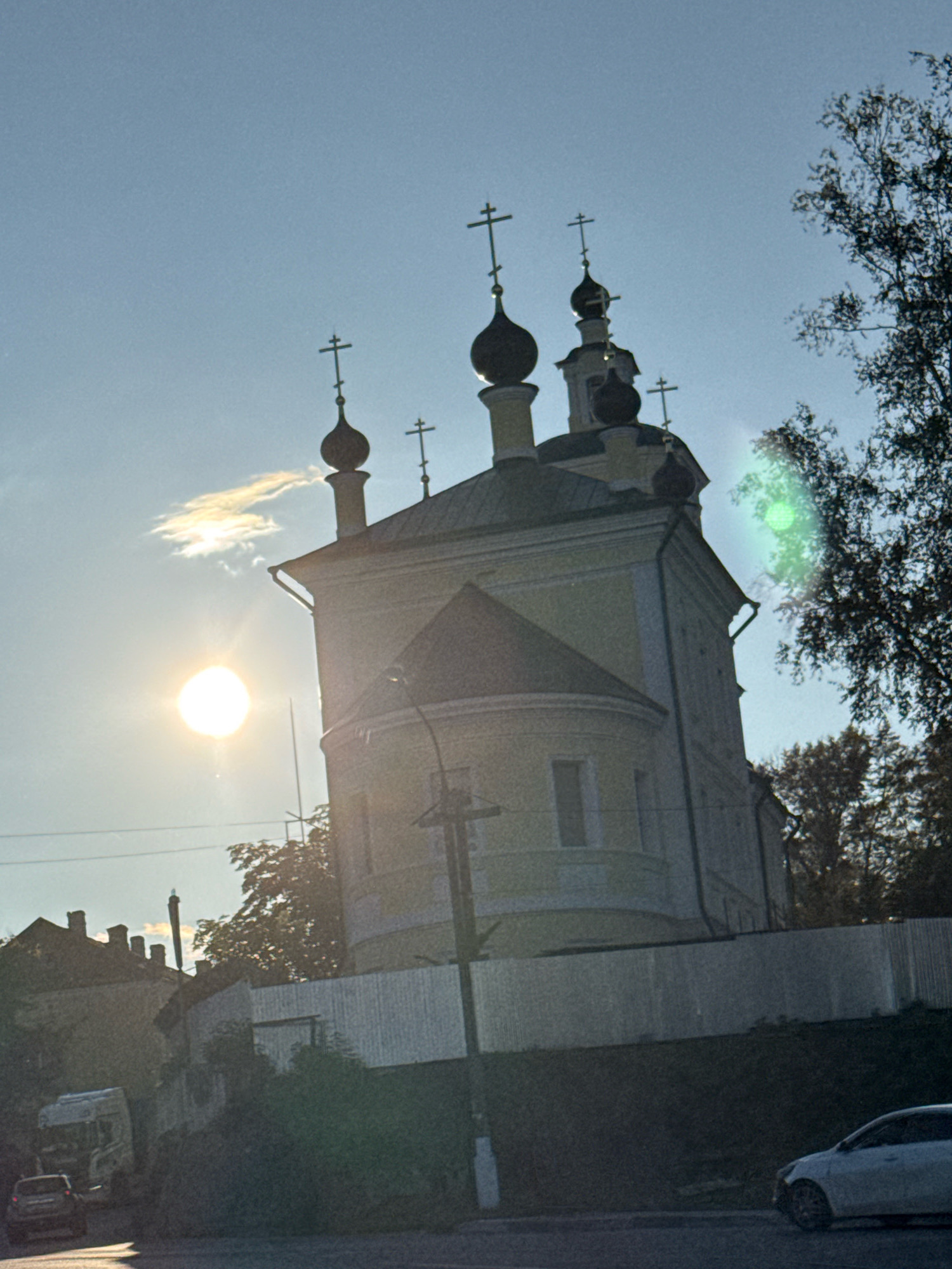 The image shows a view of the Church of St. Nicholas, located in the city of Torzhok, Russia. The church, with its distinctive yellow facade and dark onion domes topped with crosses, is silhouetted against a bright, hazy sky. The sun is prominently visible in the left portion of the frame, casting a glare that creates lens flare effects. To the right of the church, trees with dark foliage are visible. In the foreground, there is a white fence running horizontally across the image. Below the fence, a dark embankment leads down to a road where two vehicles are present: a white truck on the left and a white car on the right. The overall scene suggests an outdoor setting, likely during daylight hours, with a focus on the architectural features of the church.