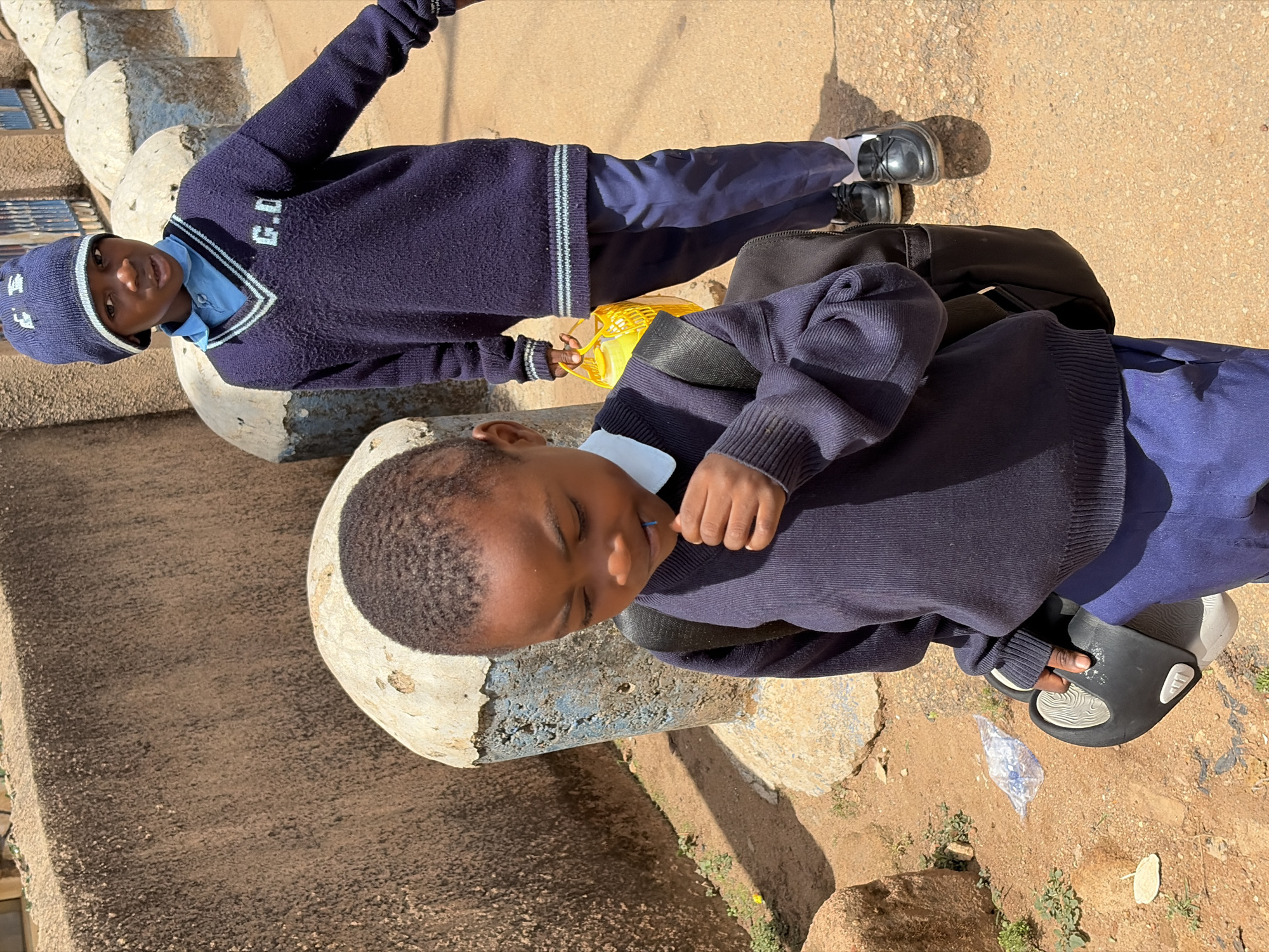 This media file features two school children, likely boys, standing outdoors on a sunny day in Jos, Nigeria. Both are dressed in dark blue school uniforms, consisting of sweaters, light blue shirts, and dark blue trousers.
The child positioned on the left wears a dark blue V-neck sweater with white and light blue stripes around the collar. The letters "G.D" are visibly embroidered in white on the left chest of the sweater. He also wears a matching dark blue beanie with white stripes and what appears to be "G D" or similar text. He looks directly at the camera with a neutral expression and holds a yellow plastic item, possibly a container or bag, in his left hand. His feet are visible, clad in black lace-up shoes and white socks.
The second child, on the right, is seen from a slightly elevated perspective. He wears a dark blue round-neck sweater over a light blue shirt, with a black backpack slung over his shoulders. His eyes are either closed or directed downwards, and one hand is held near his mouth. He wears black open-toed sandals with a distinct white, textured sole.
The scene is set on a rough, light brown concrete surface mixed with patches of dirt, small green plants, and scattered rocks. The presence of strong shadows indicates bright daylight and clear weather. A partially visible, light-colored concrete structure with some light blue paint is in the background. The children appear to be either on their way to or returning from school, given their attire and backpacks.