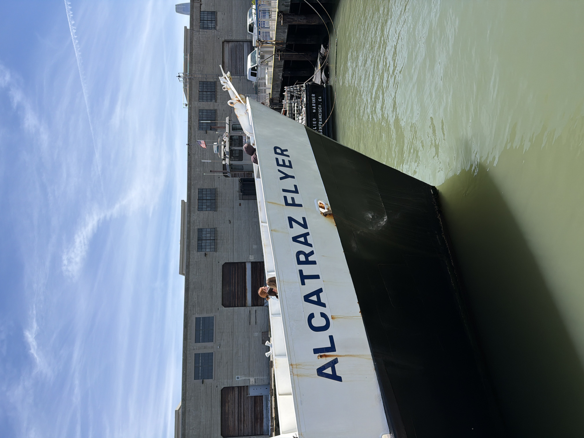 The image shows the bow of a ferry named "ALCATRAZ FLYER" docked in the waters of San Francisco, California, United States of America. The ferry, painted white and black, is positioned at an angle, with the name clearly visible in blue lettering on its side. Several people are visible on the deck of the ferry, including a woman with red hair and another person with dark hair. In the background, a gray industrial building with large windows and garage doors lines the waterfront. Adjacent to the ferry, another boat is docked, bearing the name "APPLIED MARINER SAN FRANCISCO, CA" in white lettering. Vehicles, including white vans, are parked on a pier behind the boats. The sky above is a clear blue with wispy white clouds and a contrail. The water in the foreground is a murky green color.