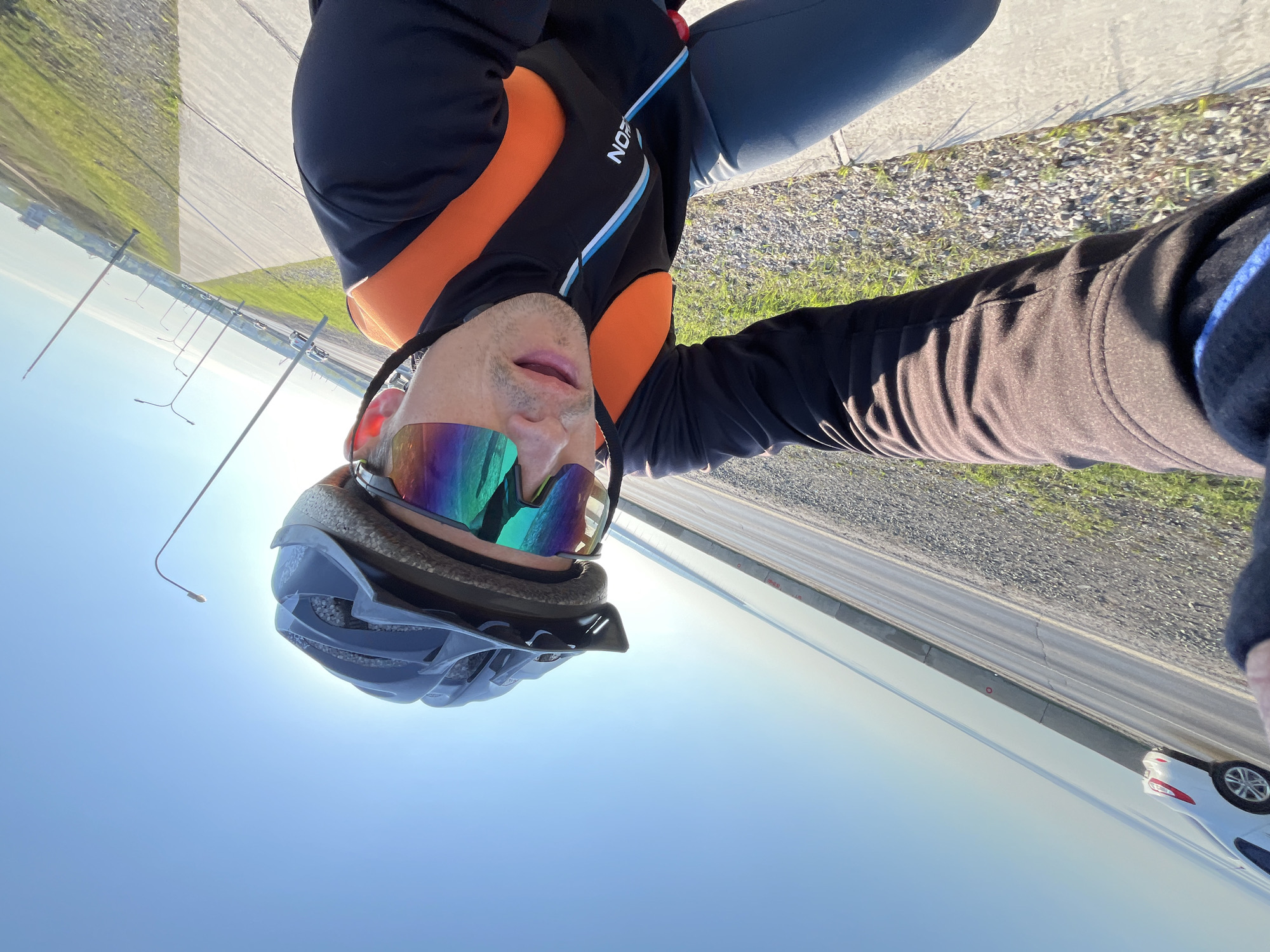 A selfie depicts a man wearing a blue helmet and rainbow-tinted sunglasses, positioned in what appears to be a roadside environment. The man is wearing a black jacket with orange panels and blue accents. The photo includes elements of an outdoor setting, such as a road with visible road markings, grass, street lights and vehicles. Based on the presence of the street lights and cars, and the style of road, the location might be a road or highway in the vicinity of Chaikovsky, Russia. The angle of the shot indicates the man may be on a bike and taking a selfie.