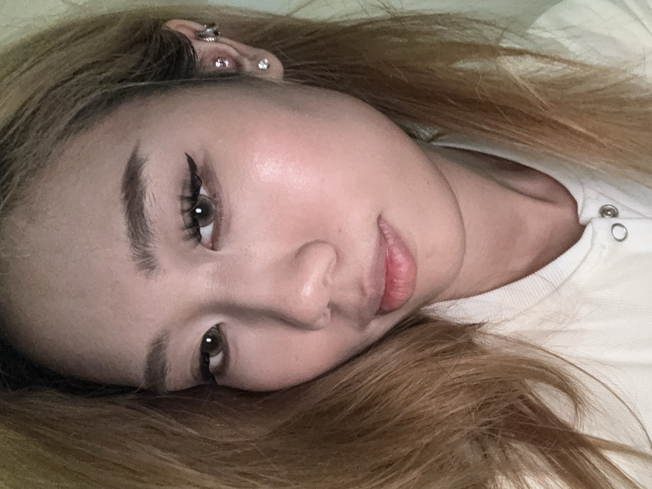 A close-up, frontal portrait displays a single female individual, located in Yuzhong District, China. The subject, appearing to be a young adult, possesses fair skin and light brown hair that frames her face. Her eyes are brown, enhanced with winged eyeliner and extended eyelashes. Her eyebrows are well-defined. Visible makeup includes a subtle cheek highlight and tinted lips. The individual wears a light-colored, possibly off-white, collared garment featuring a line of silver-colored snap buttons down the front. The subject's visible right ear has a single stud earring in the lobe, two additional studs in the upper cartilage, and a small silver-toned hoop earring further up the helix. She maintains a direct gaze towards the camera with a neutral facial expression. The background is a uniformly light, out-of-focus plane, suggesting an indoor setting with minimal detail.