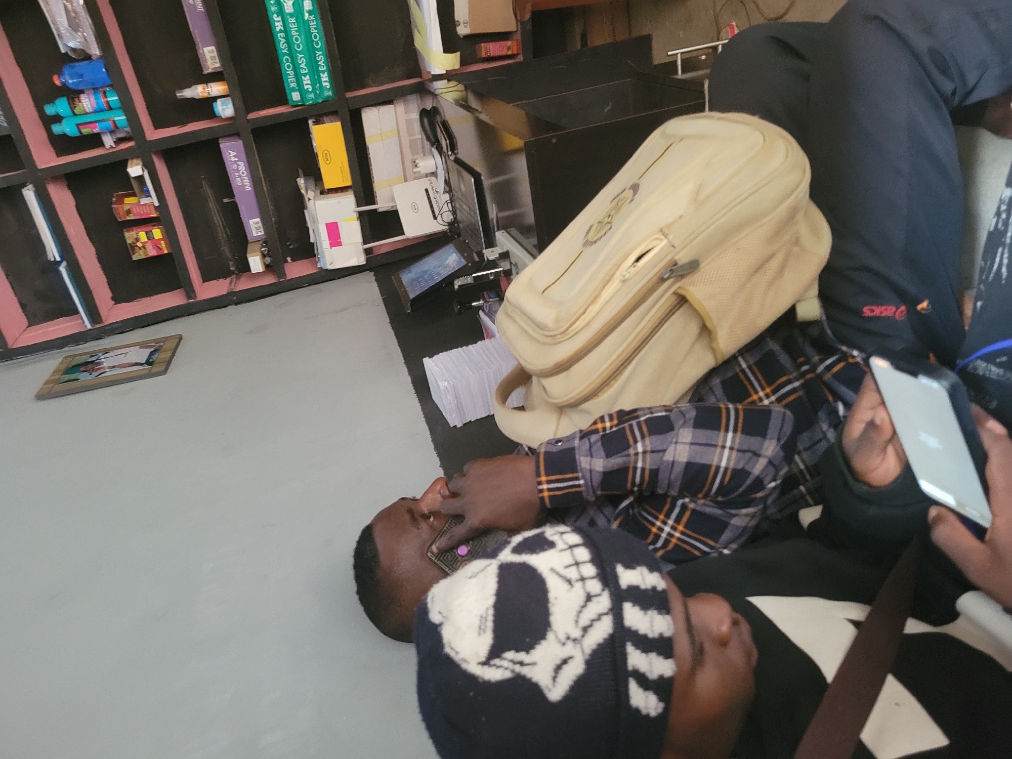 In Bokkos, Nigeria, a scene unfolds within an office or shop. Two individuals are present. The foreground features a person wearing a black beanie with a white skull design and a black sweatshirt. This individual is looking down at a silver tablet, holding it with both hands. To their right, a second person, facing to the right of the frame, is on the phone. This person is wearing a plaid shirt with orange, black, and white checkered patterns, and is holding a dark-colored phone to their right ear. A tan messenger bag rests on their lap, draped over their knees, with a portion of the bag visible.
The environment includes a desk area behind the individuals. On the desk, a laptop is open and powered on, displaying a screen with a blue background and graphical elements. A Wi-Fi router, a black phone, a stapler, and other office supplies are also on the desk. Behind the desk, shelves are filled with various items, including stacks of paper, boxes, bottles of cleaning supplies, and what appear to be reams of copier paper. A framed picture is visible on the wall to the left of the shelves. The lighting suggests an indoor setting, possibly illuminated by overhead lights or natural light from an unseen source. The overall impression is of a functional, perhaps slightly cluttered, workspace.
