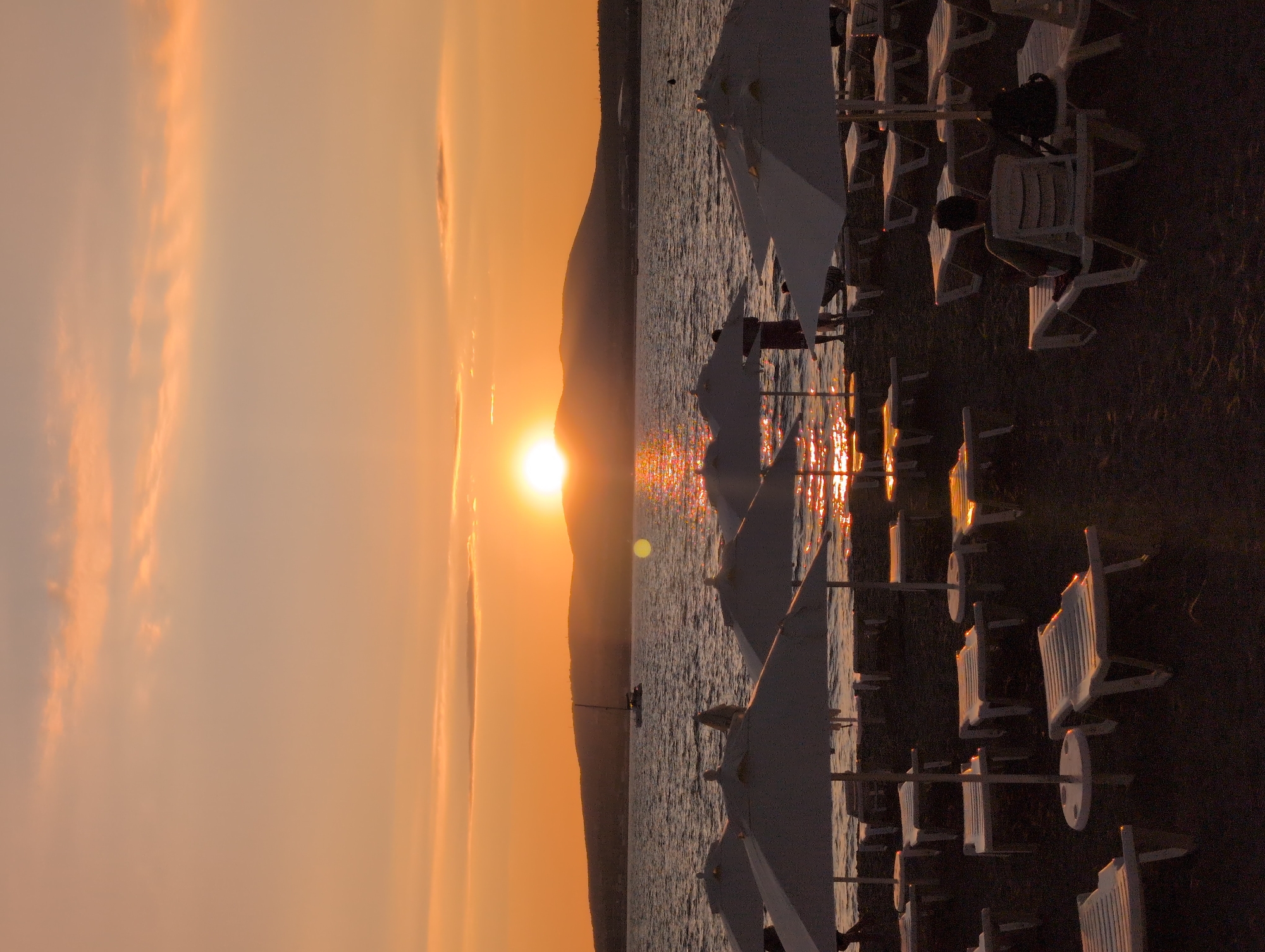 A serene sunset paints the sky over the Black Sea in Gelendzhik, Russia. The sun, a brilliant orb of light, dips behind a distant mountain range, casting a warm, orange glow across the water. Numerous white beach umbrellas and empty lounge chairs dot the sandy shore, suggesting a leisurely afternoon that has transitioned into evening. The gentle waves of the sea reflect the sunset's golden light, creating a shimmering path across its surface. A lone sailboat glides across the water in the distance, adding to the tranquil atmosphere. A few people are visible on the beach, some standing and others relaxing on the lounge chairs, their figures silhouetted against the vibrant sky. The scene evokes a sense of peace and relaxation, capturing the beauty of a coastal sunset.