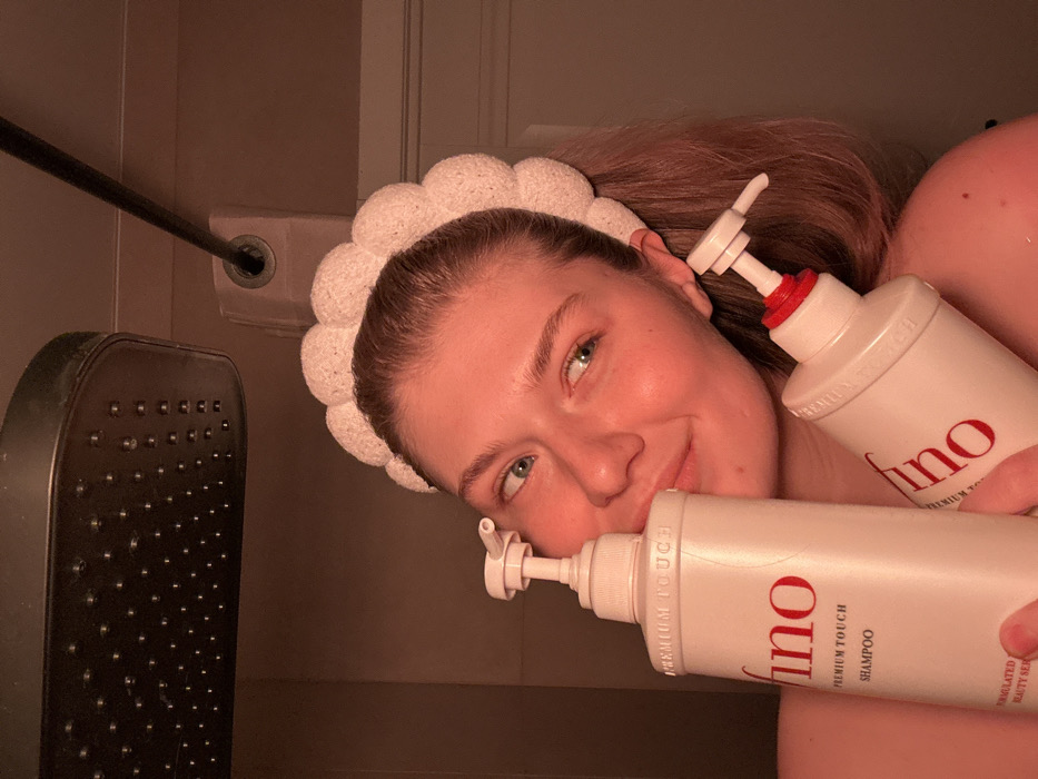 A light-skinned woman with blue eyes smiles, tilting her head. She wears a white, fluffy headband. She holds two large, white bottles of "Fino Premium Touch Shampoo." A black showerhead and shower rail are visible to the left. The overall lighting is warm and slightly dim.
