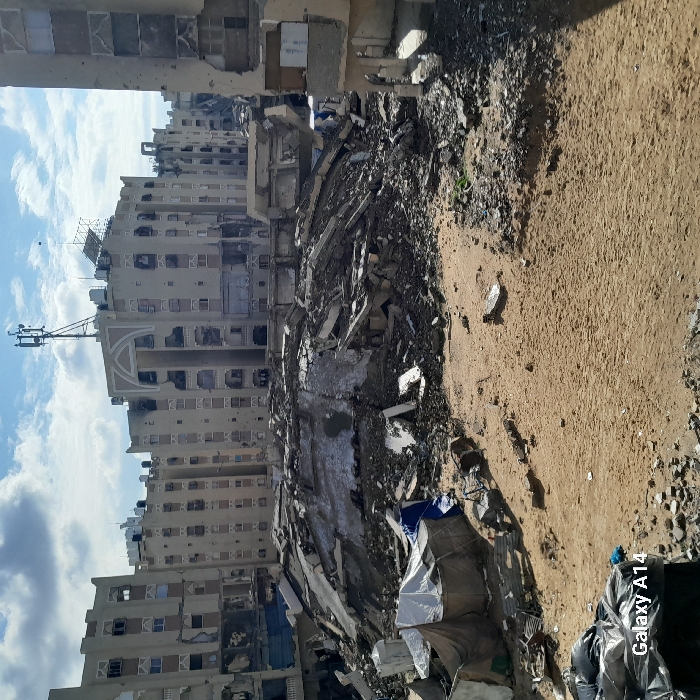The image depicts an extensively damaged urban landscape in Khan Yunis, Palestinian Territory, under a partially cloudy blue sky. The foreground consists of uneven, sandy-colored ground, sparsely littered with small debris, along with a pile of dark objects, possibly refuse bags, in the lower left corner. Adjacent to this, a makeshift shelter constructed from white and blue tarpaulins or fabric is visible. The midground is dominated by a massive mound of concrete rubble, twisted rebar, and fragmented building materials, indicative of a completely collapsed structure; some sections of this debris appear dark and possibly wet.
In the background, multiple multi-story buildings, likely residential or commercial, stand severely damaged. These structures exhibit numerous broken or missing windows, impact damage to exterior walls, visible scorch marks, and partially collapsed sections. A prominent building in the central background features distinctive arched architectural elements on its upper floors and supports a cellular transmission tower on its roof, alongside several black water tanks. The overall scene indicates widespread destruction, with no individuals or active events discernible.