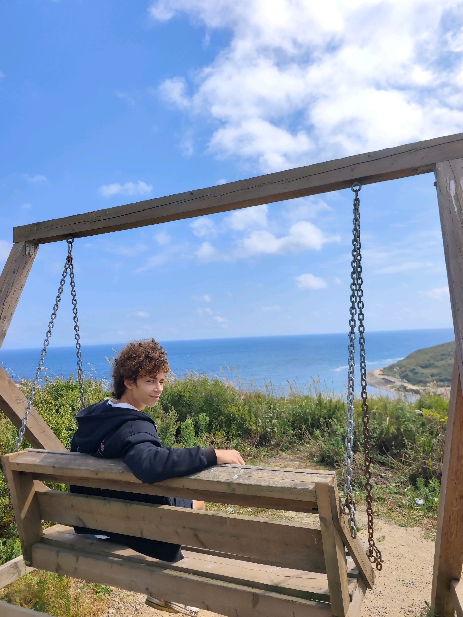 A young person with curly brown hair is sitting on a wooden swing overlooking the sea. They are wearing a black hoodie and dark pants. The swing is suspended from a wooden frame and hangs over a grassy slope leading down to a beach and the ocean. The sky is blue with scattered white clouds. The scene appears to be outdoors in a scenic coastal location. This location is not identified as being in the city of Russky, Russia.