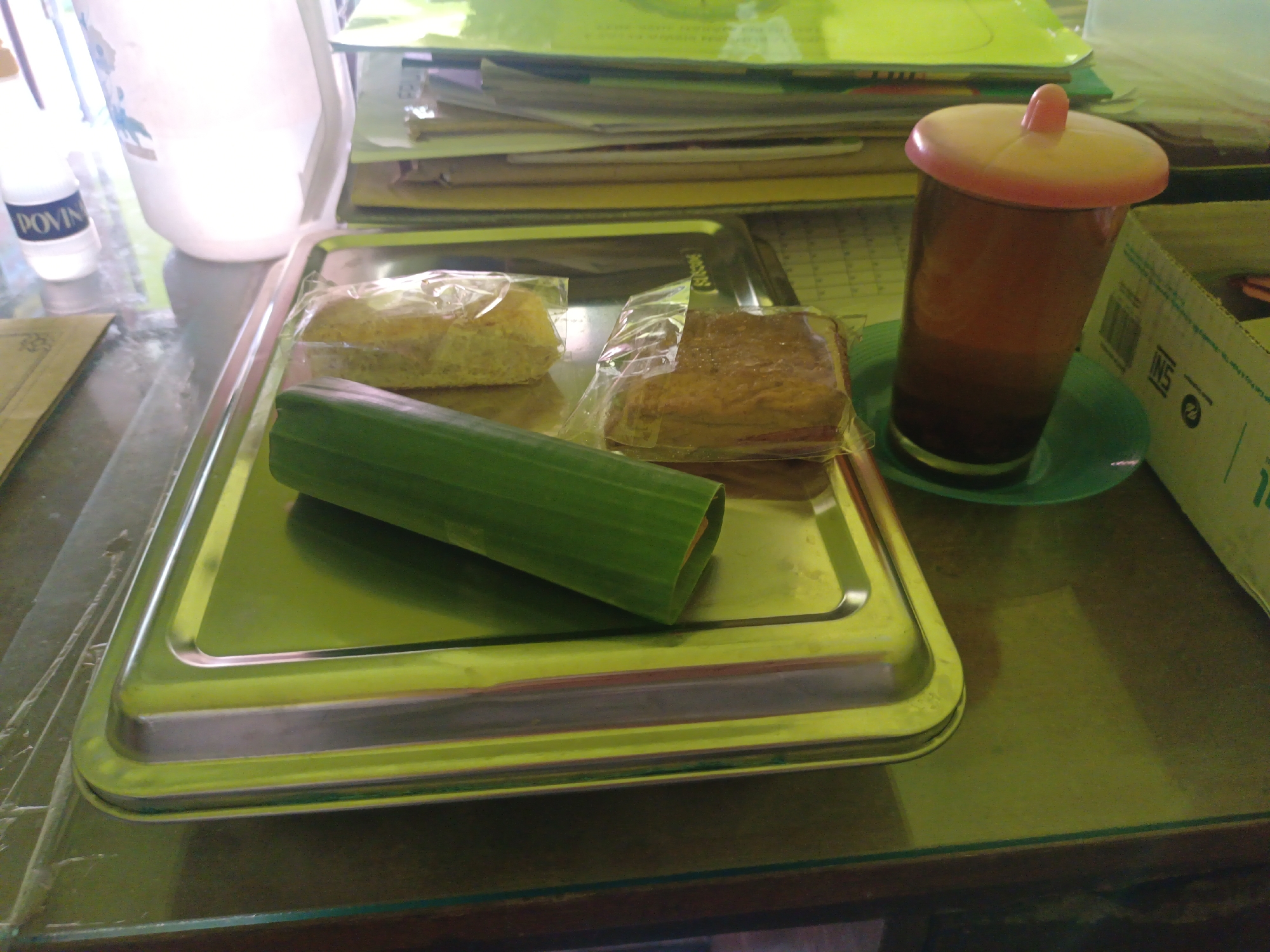 A close-up, eye-level shot displays a reflective metal tray containing two food items wrapped in plastic and a green cylindrical object. The food items appear to be some kind of cake or bread. The green object is smooth and elongated. To the right of the tray, a transparent glass with a brown beverage and a pink lid sits on a green saucer. In the background, a stack of papers and a partially visible white container are present. The scene is set on a glassy surface, likely a table, with a hint of a wooden surface below. The overall lighting is dim, creating a warm, amber hue. The image is captured at a residence in Sarimulyo, Indonesia, suggesting a domestic setting.